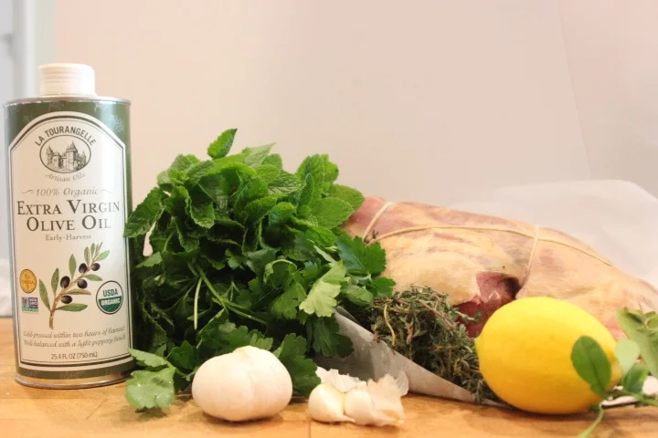A bunch of fresh cilantro, a garlic bulb with cloves, a lemon, a bundle of thyme, a whole pork roast, and a bottle of extra virgin olive oil on a wooden surface.