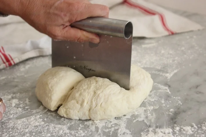 A person using a bench scraper to cut dough on a floured surface in a kitchen.