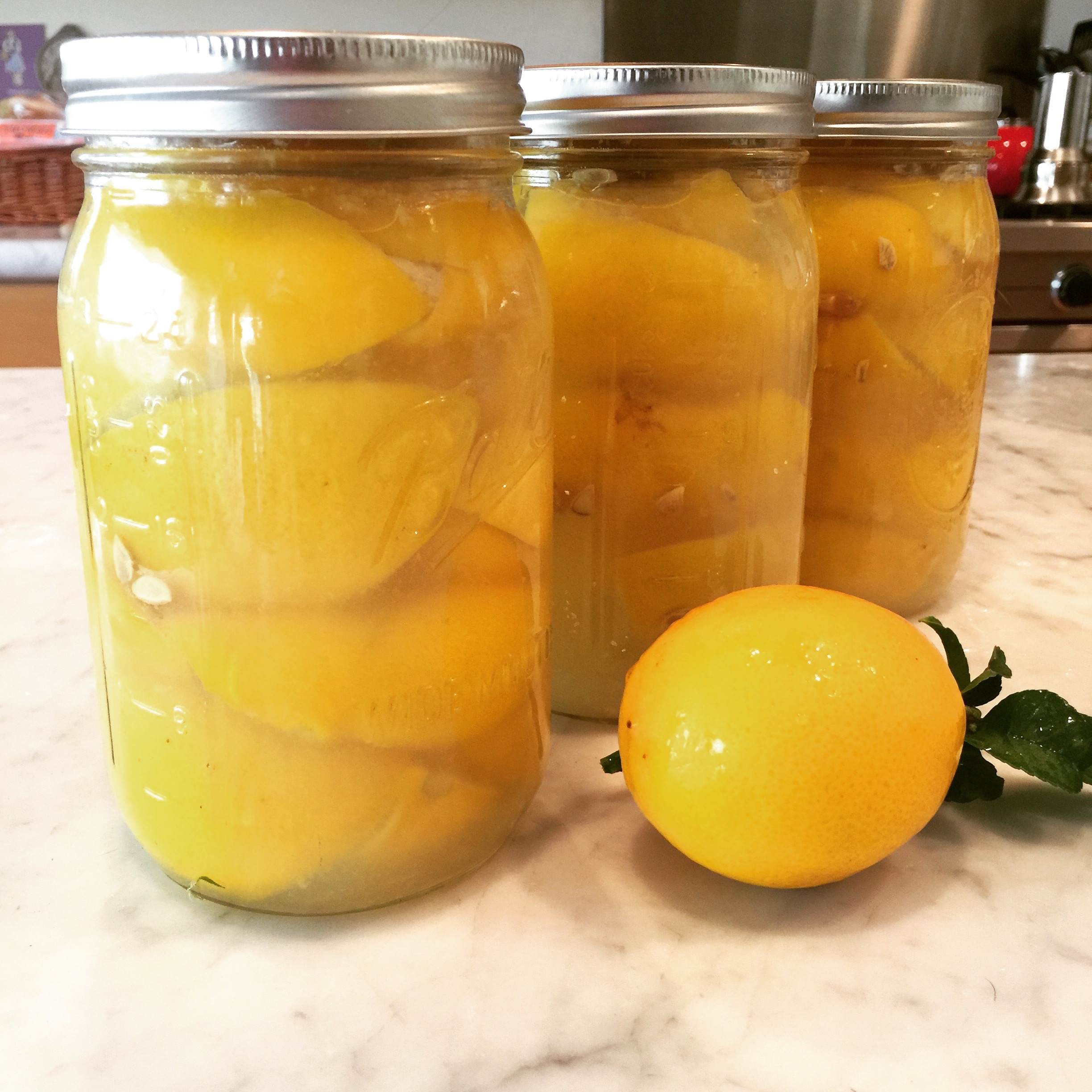 Three mason jars filled with preserved peaches and peach slices are displayed on a kitchen counter. An orange peach with green leaves is placed in front of the jars.