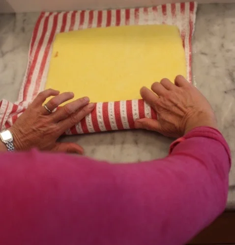 Hands wrapping cheese in a red and white striped cloth on a marble surface.