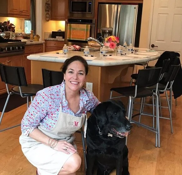 A woman kneeling next to a black dog in a kitchen with a restaurant-style apron, smiling at the camera.