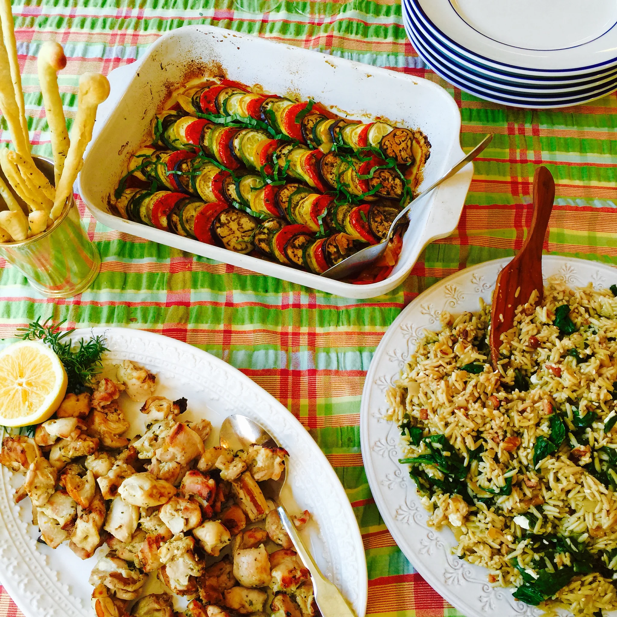 A colorful table with a large white dish of layered grilled vegetables, a plate of cooked rice with vegetables, and other dishes and lemon slices.