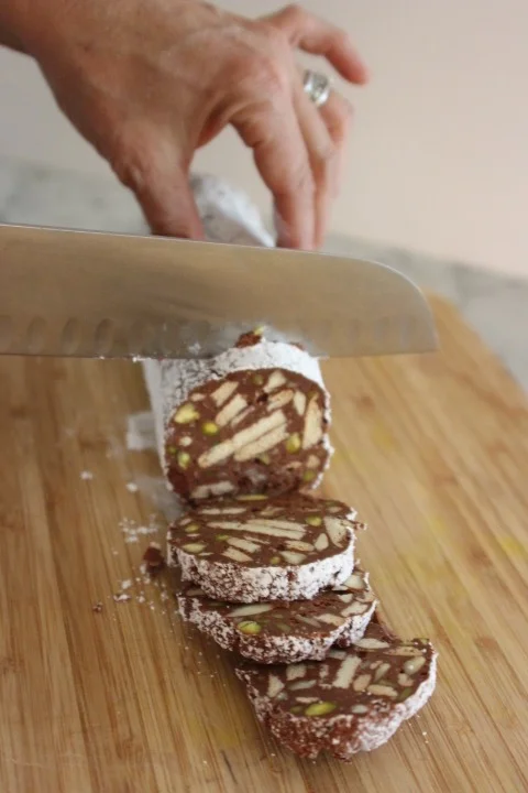 A hand is slicing a rolled chocolate confection with nuts and pistachios on a wooden cutting board.