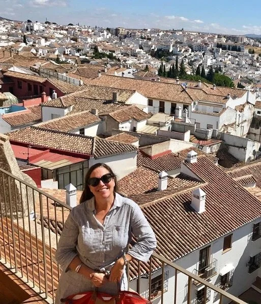 Woman standing on a terrace with a cityscape of white buildings and red-tiled rooftops in the background, wearing sunglasses and a gray shirt.