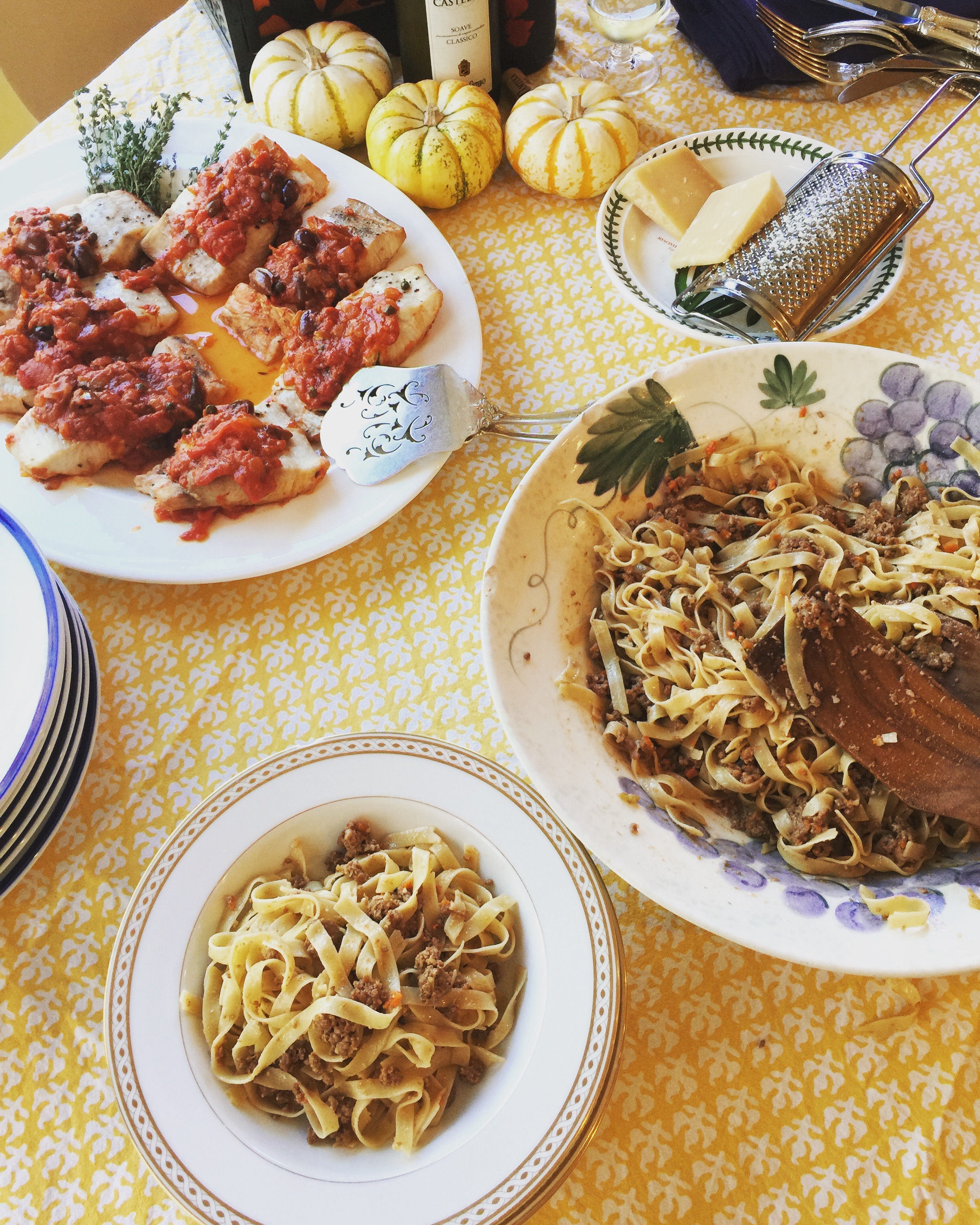 Plate of cooked fettuccine with meat sauce on a yellow and white patterned tablecloth, accompanied by small decorative pumpkins, cheese, a grater, and glasses of cream or sauce.