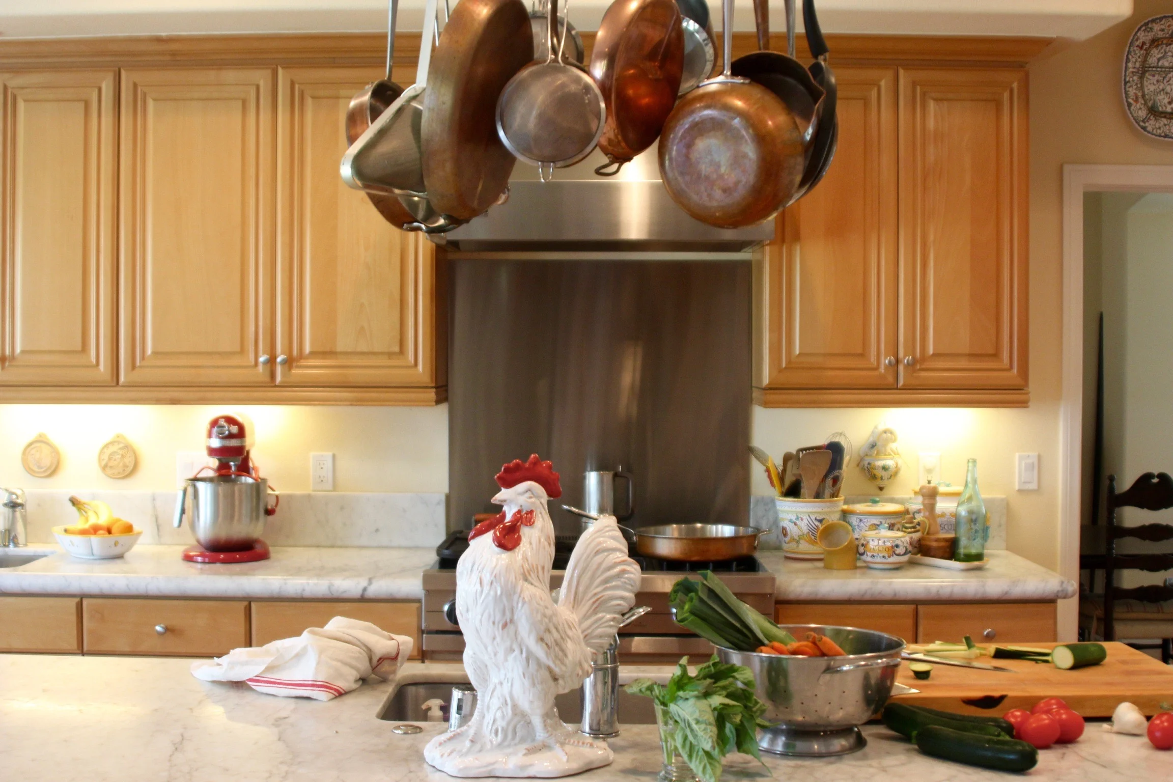 Kitchen with hanging copper pots and pans above a stove, decorated with rooster figurines on the counter, surrounded by vegetables and kitchen utensils.