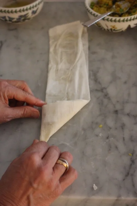 A person folding a piece of parchment paper, with bowls of food in the background on a marble surface.