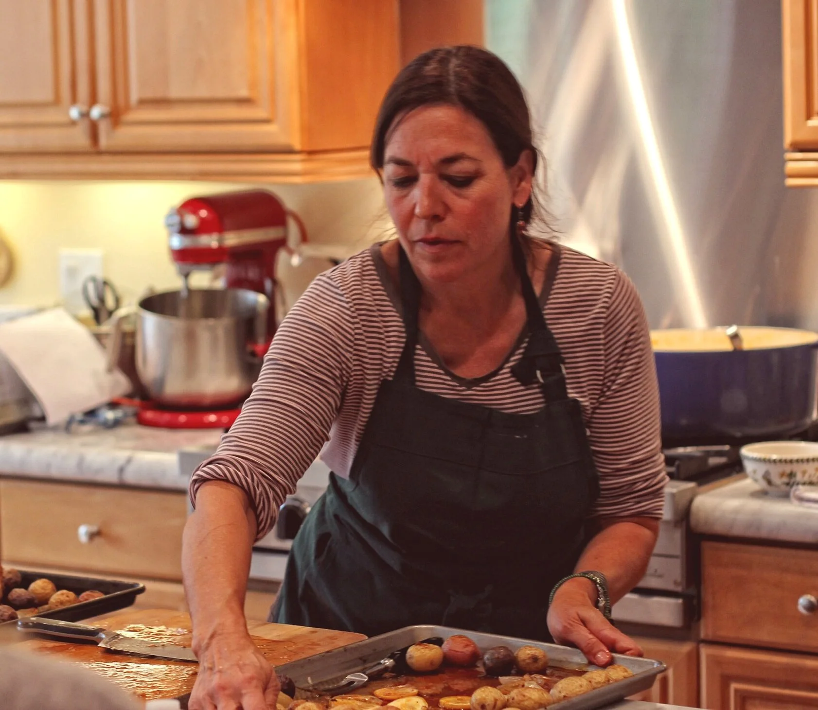A woman wearing a black apron and a striped shirt is preparing food in a kitchen. She is working on a tray filled with small round vegetables or potatoes. The kitchen has wooden cabinets, and a red stand mixer is visible in the background on the coun