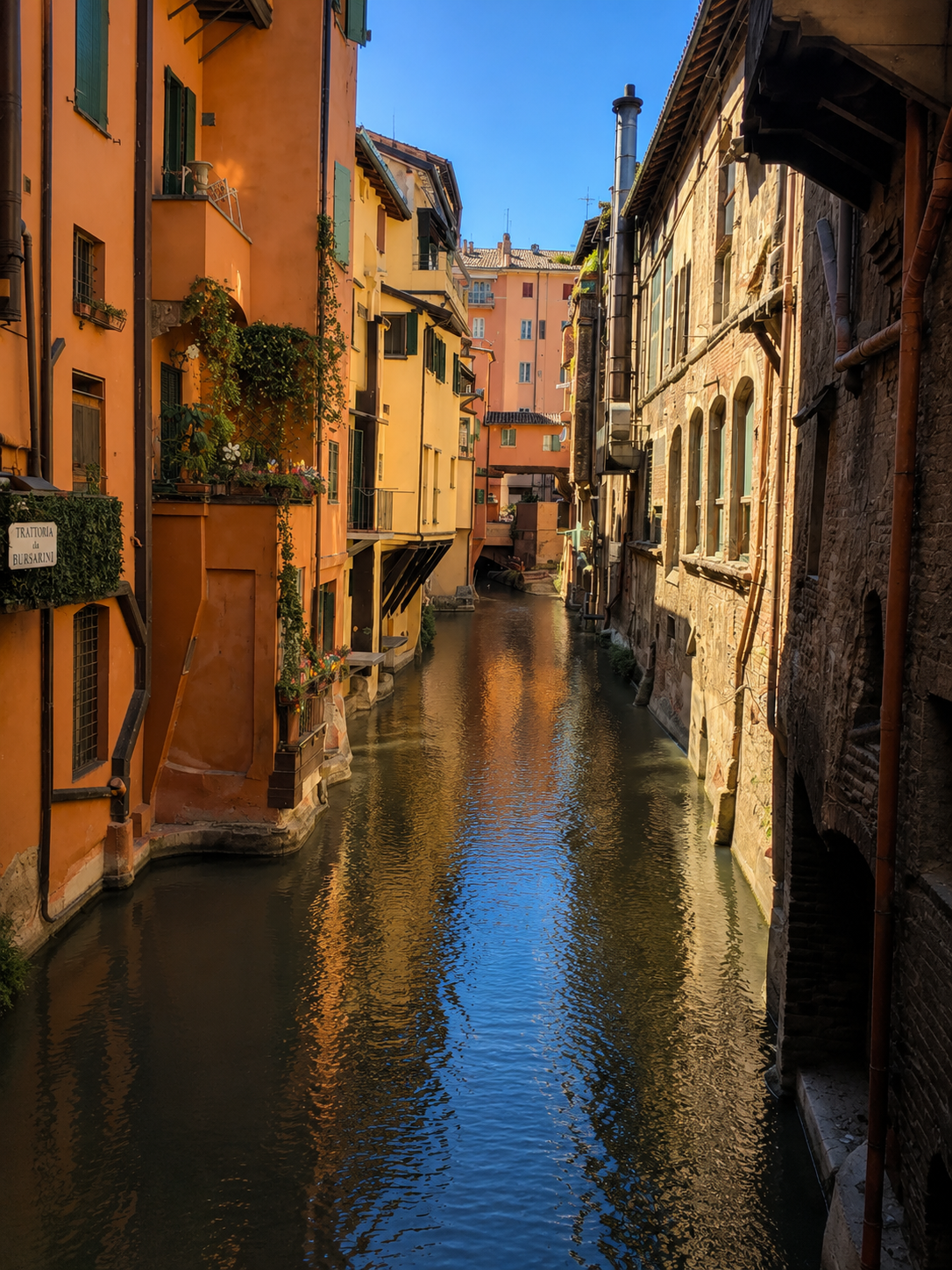 A narrow canal lined with colorful, multi-story buildings on both sides, reflecting in the water, under a clear blue sky.