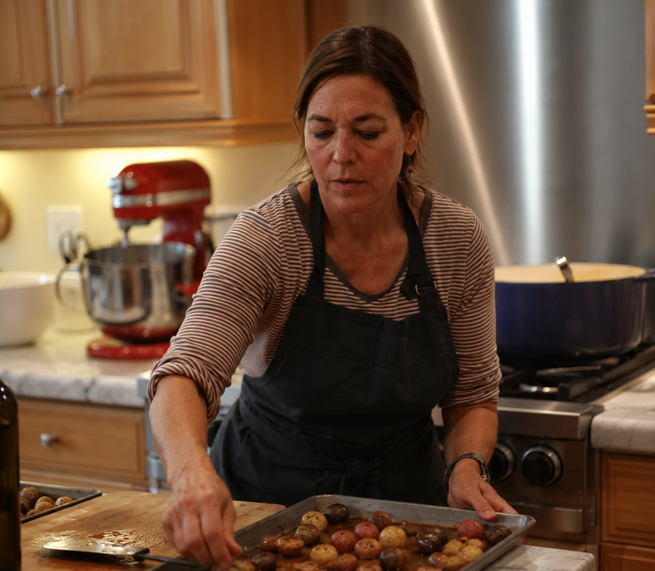 A woman wearing a black apron and striped shirt preparing a tray of roasted potatoes in a kitchen.