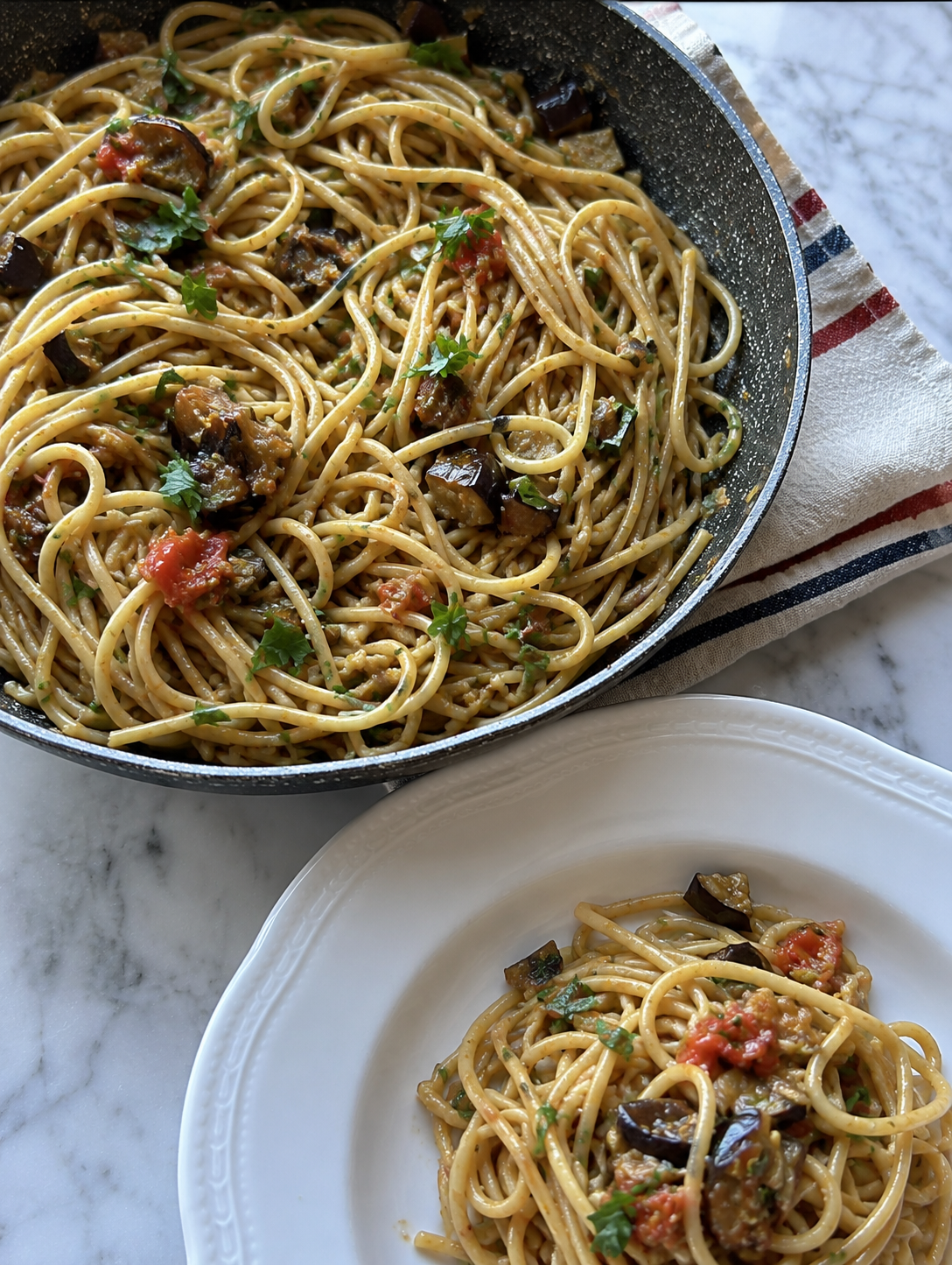 A large black skillet and a white plate both contain spaghetti pasta with tomato sauce, eggplant, and fresh parsley, on a marble surface with a striped cloth underneath.