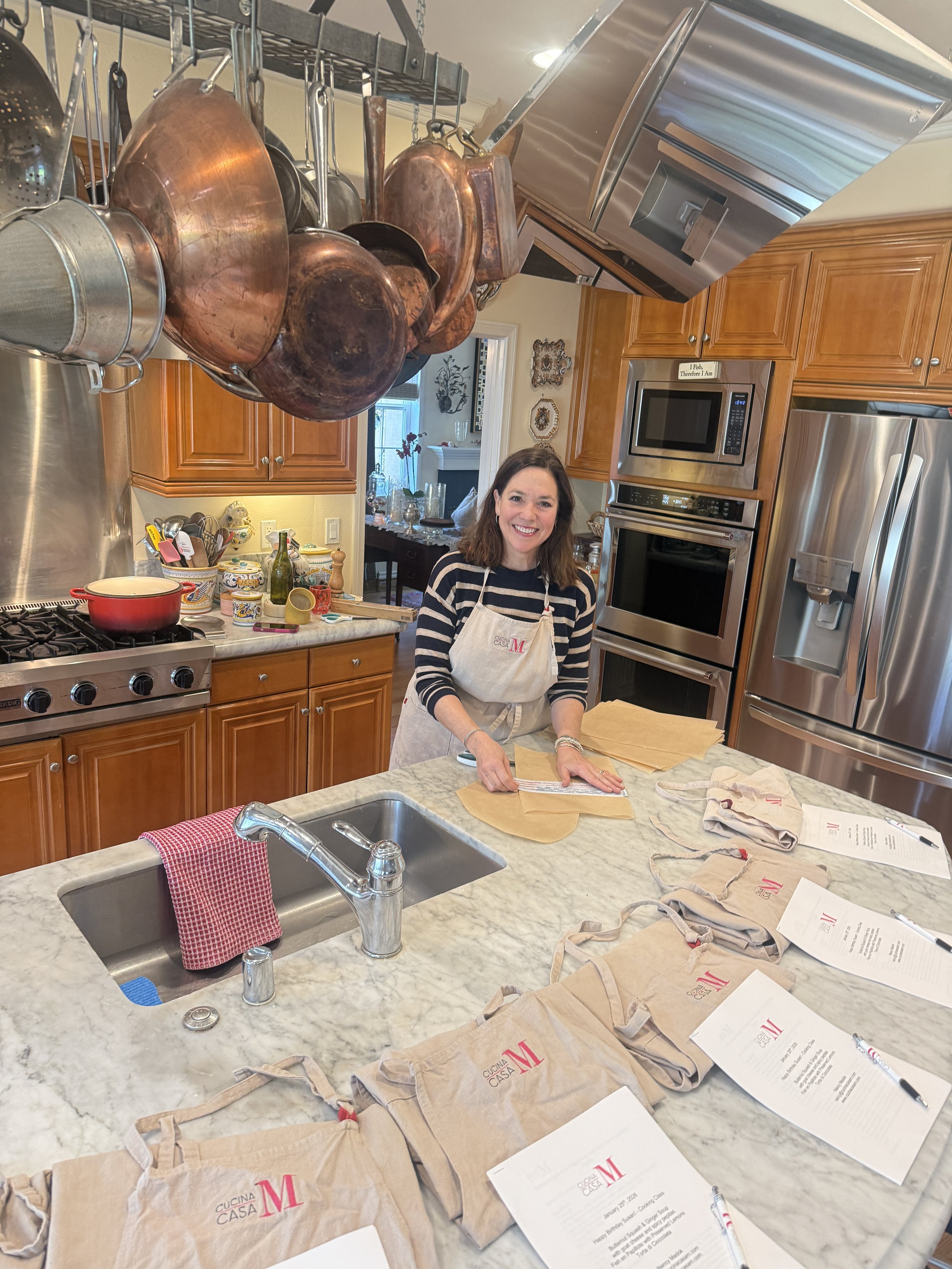 A woman in a white apron leading a cooking class with five women sitting around a kitchen island, with herbs, lemons, and cooking ingredients on the counter.