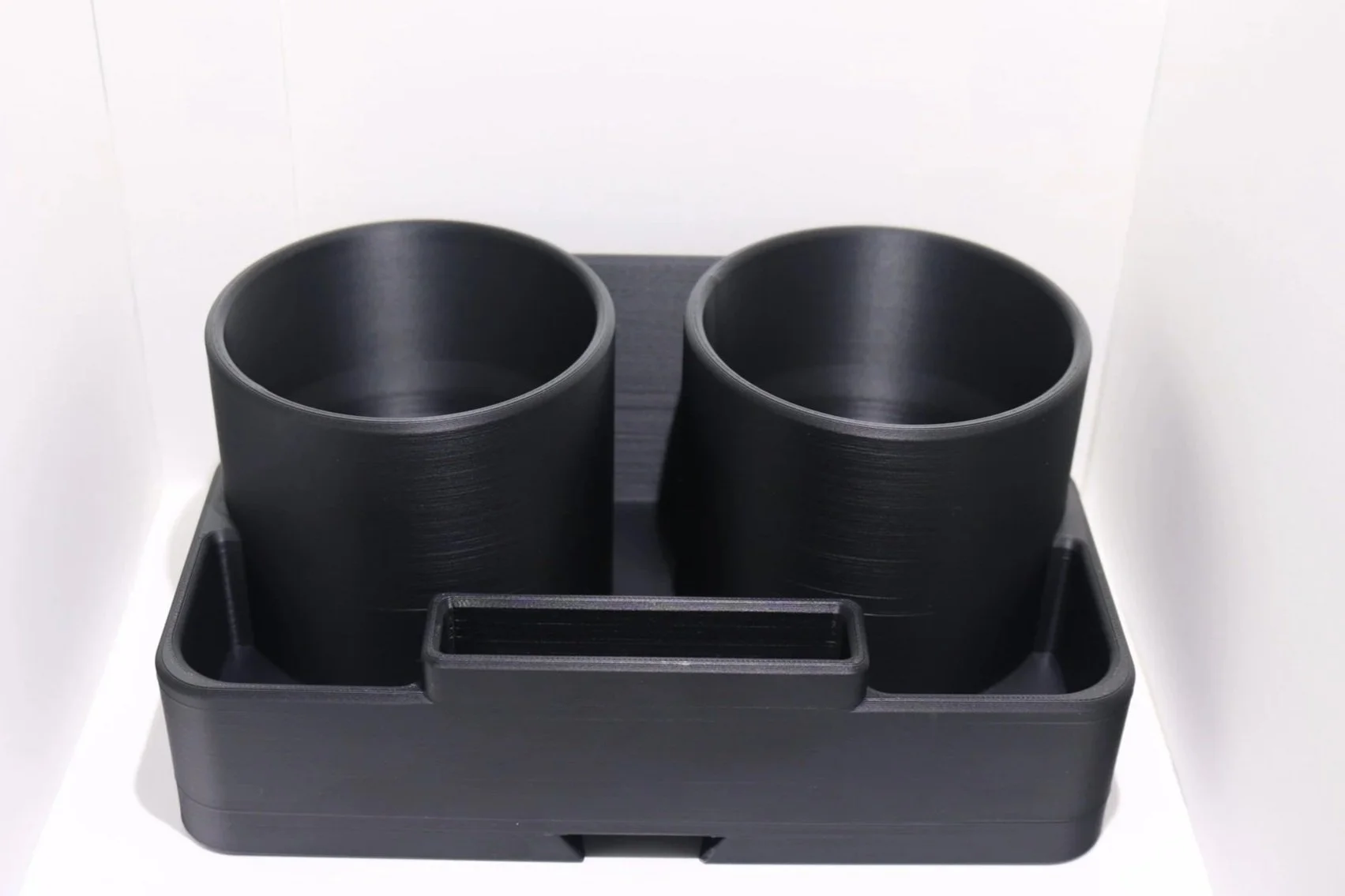 Two black plastic cylindrical cups on a black plastic base with a rectangular slot in front, against a white background.