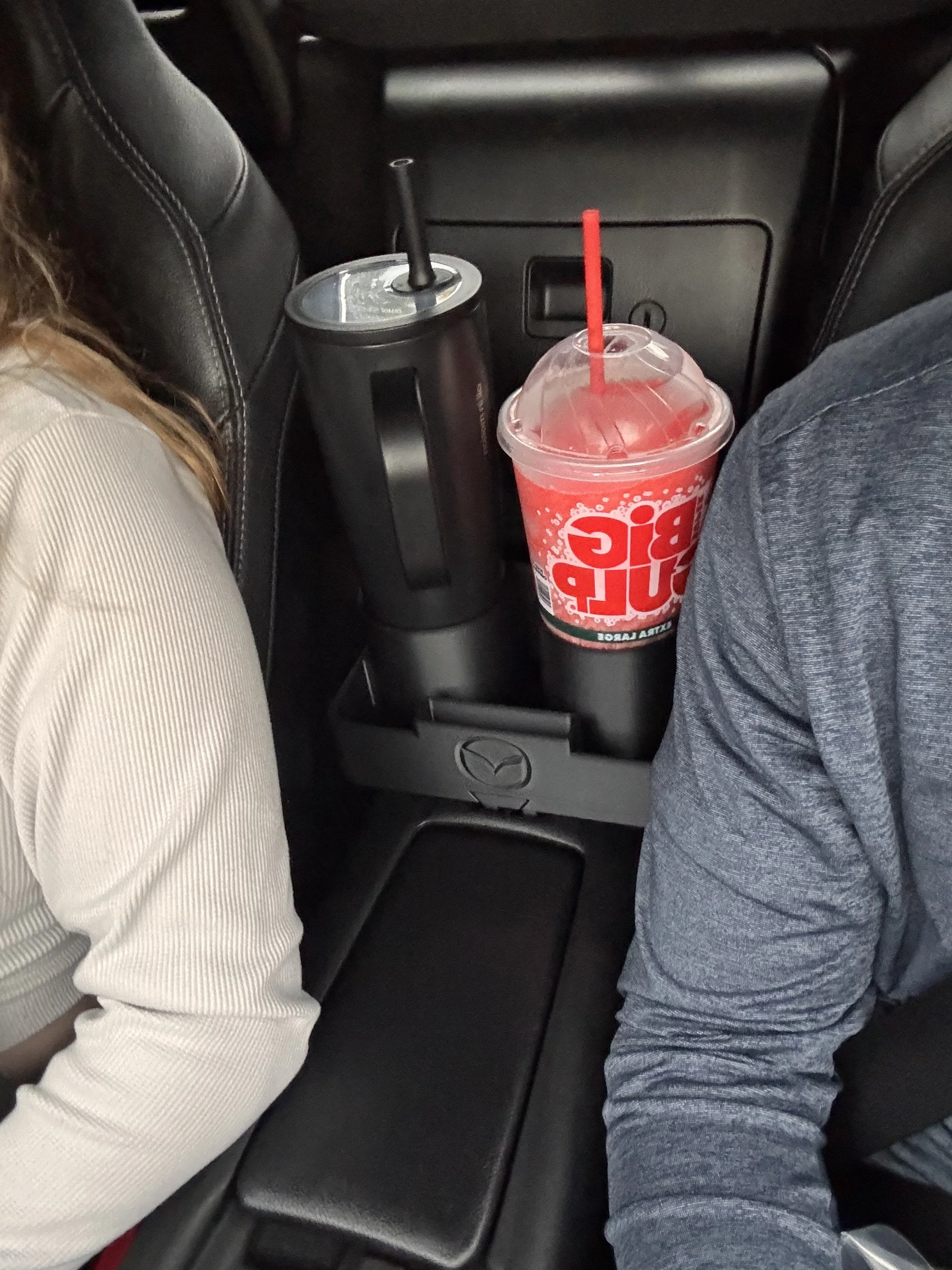 Inside the backseat of a car showing two cupholders with drinks, one with a McDonald's drink and straw, and a can in the other cupholder, between two seated passengers.