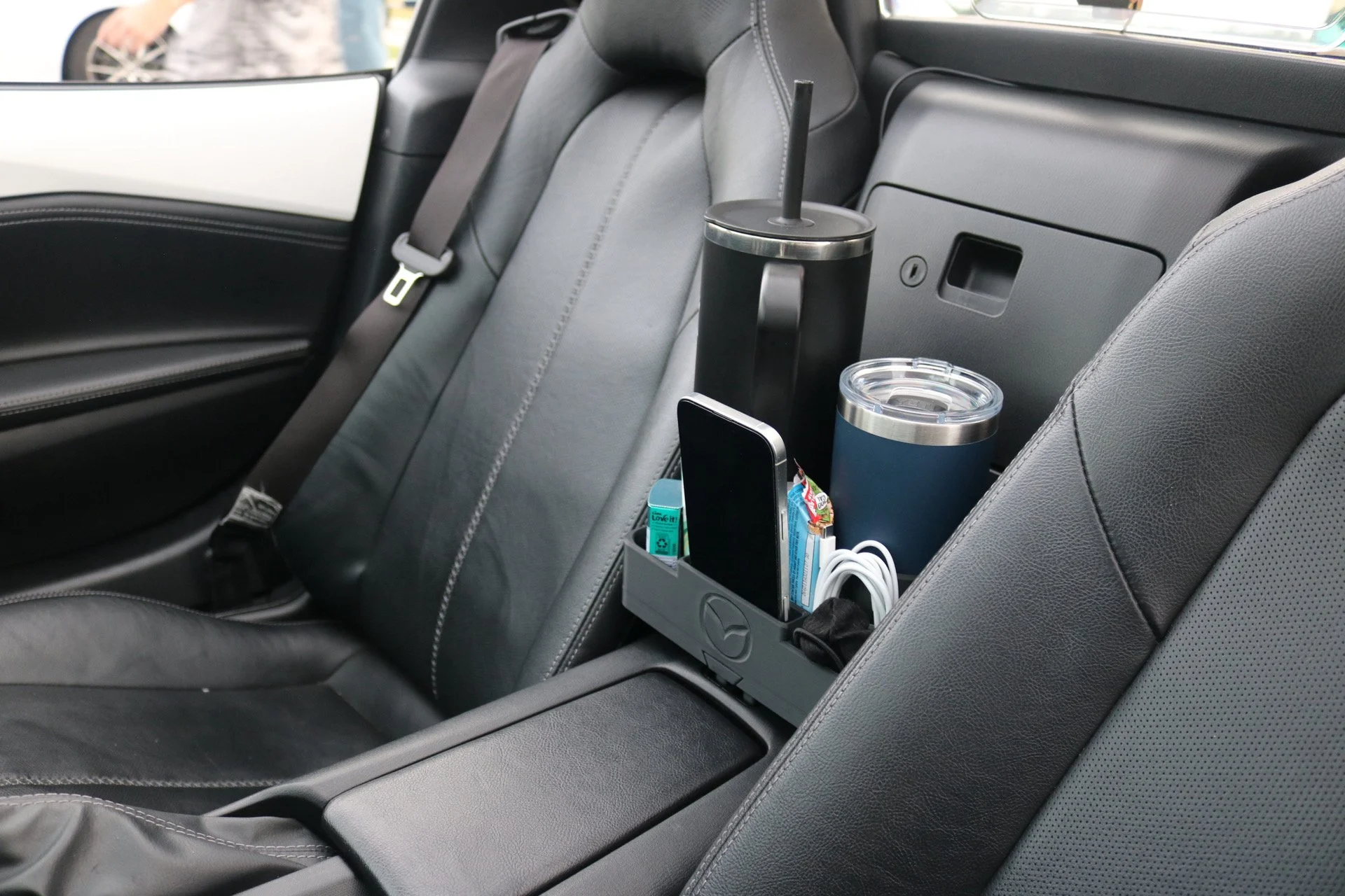 The back seat of a car with a built-in center console holding two travel mugs, a smartphone, a pack of gum, and charging cables.