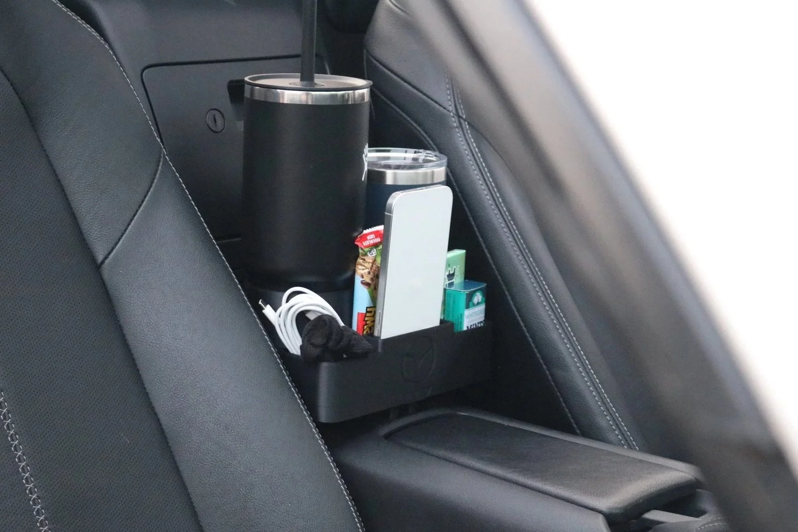 Car back seat with a seatback pocket holding a large black tumbler, a smaller silver tumbler, a white smartphone, a charger, and various small snack and medication packages.