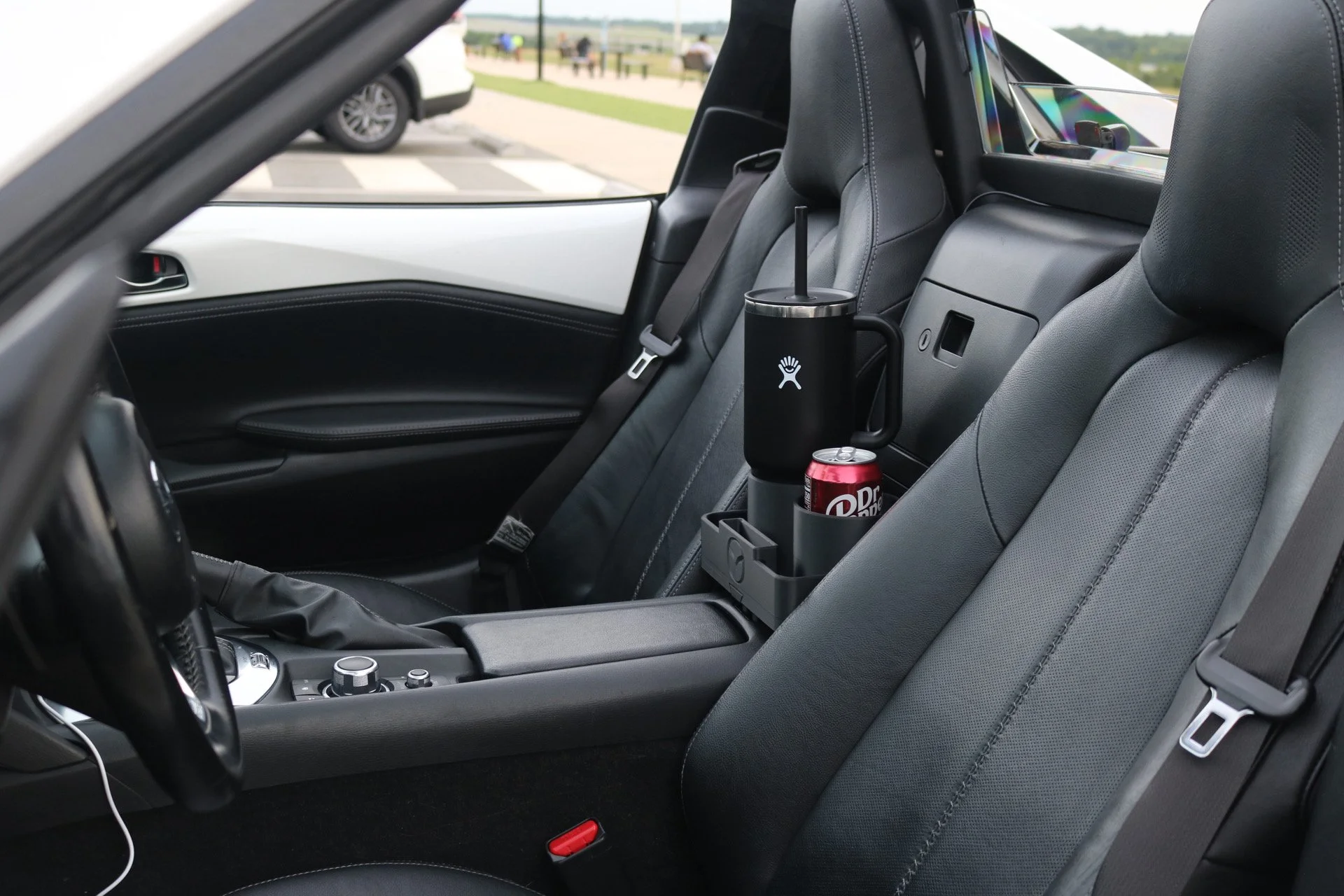 Interior of a car showing black leather seats, a center console, and a cup holder with a black tumbler and a can of Dr. Pepper, with a parking lot and people in the background.