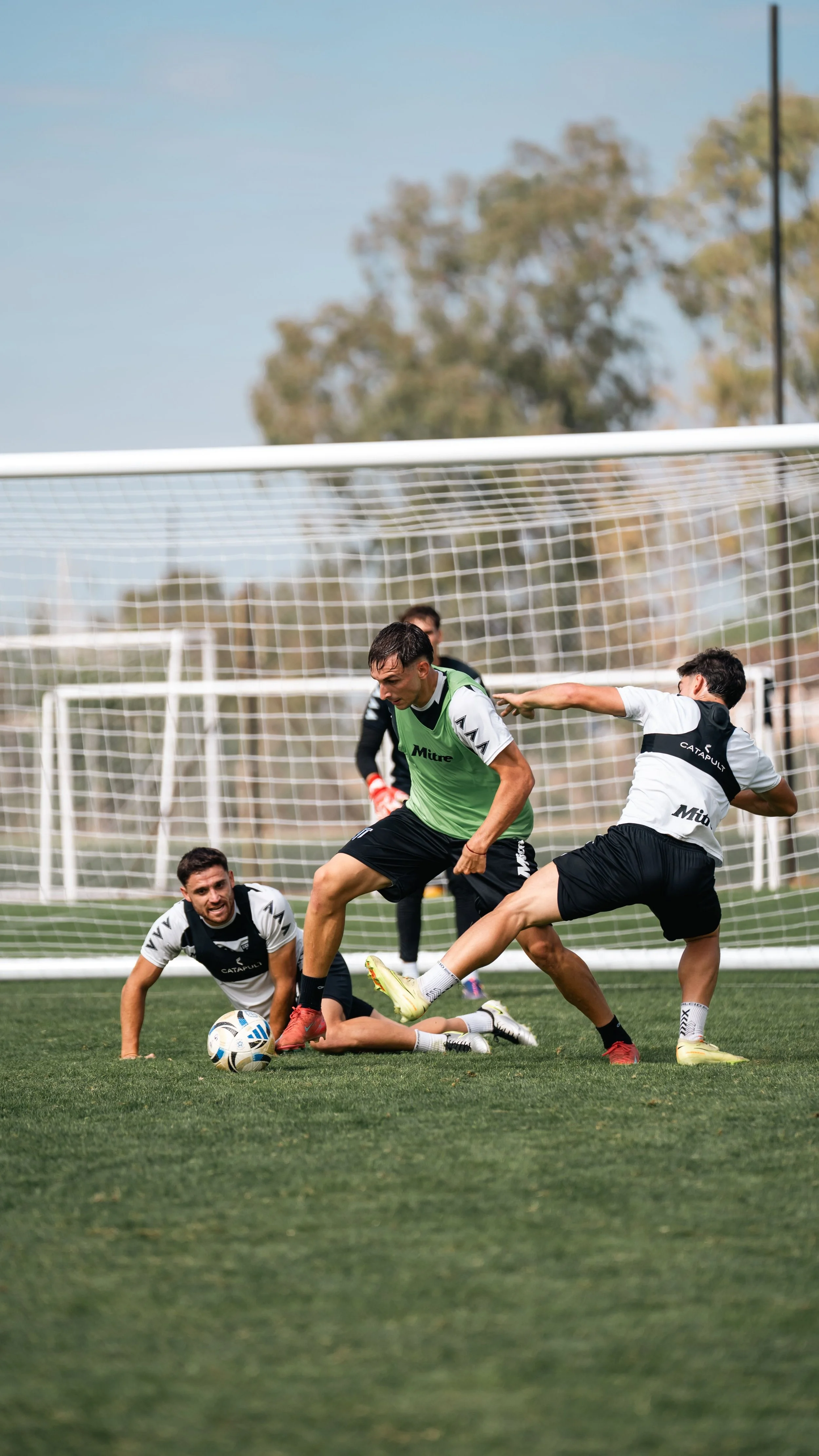 Soccer players competing for the ball near the goal on a grassy field, with a net and trees in the background.