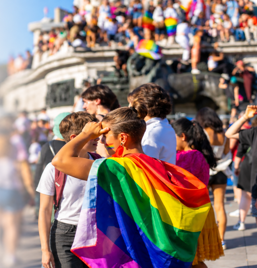 Person with rainbow pride flag draped over shoulders in front of a crowd at a pride event.