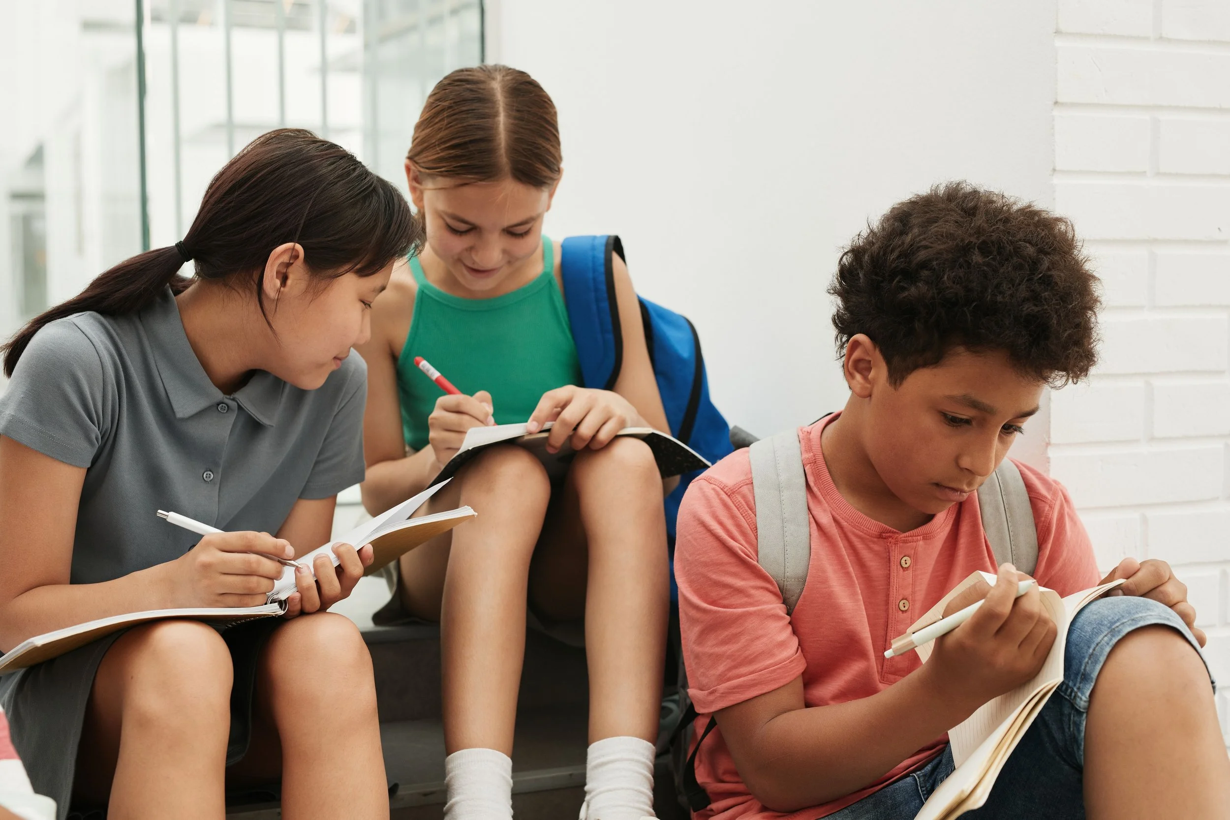 Three students sitting on stairs, reading their books and notes, with a school environment in the background.