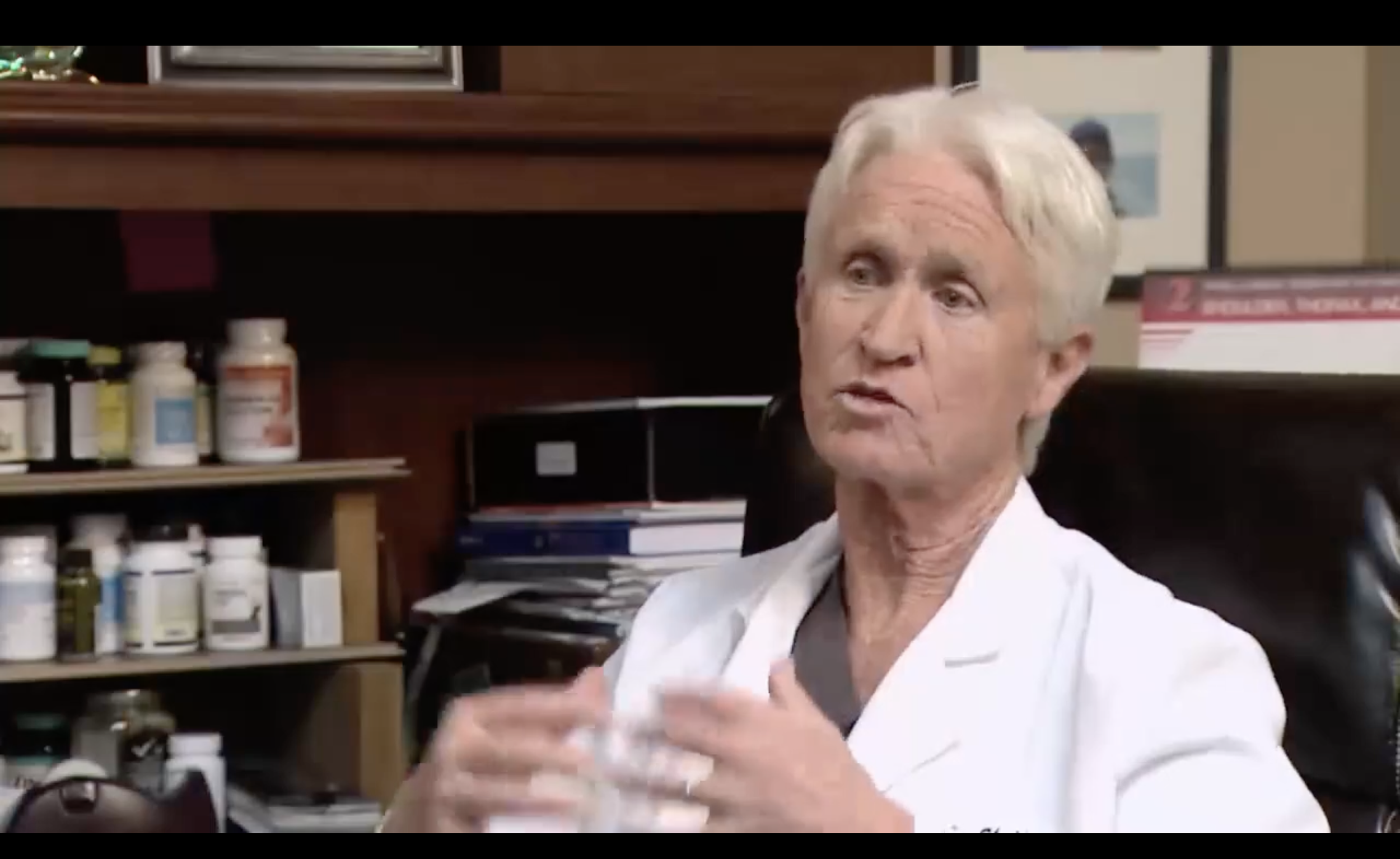 An elderly man with white hair wearing a white coat is speaking or explaining something in an office or medical setting, with shelves of medication bottles behind him.