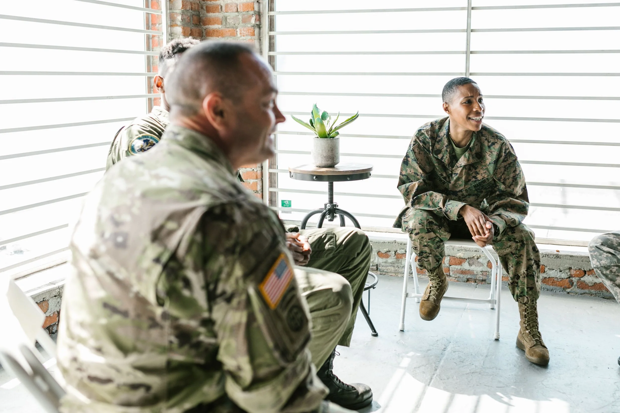 Group of military personnel sitting and having a discussion in a room with brick wall and large windows, with a woman smiling and talking.