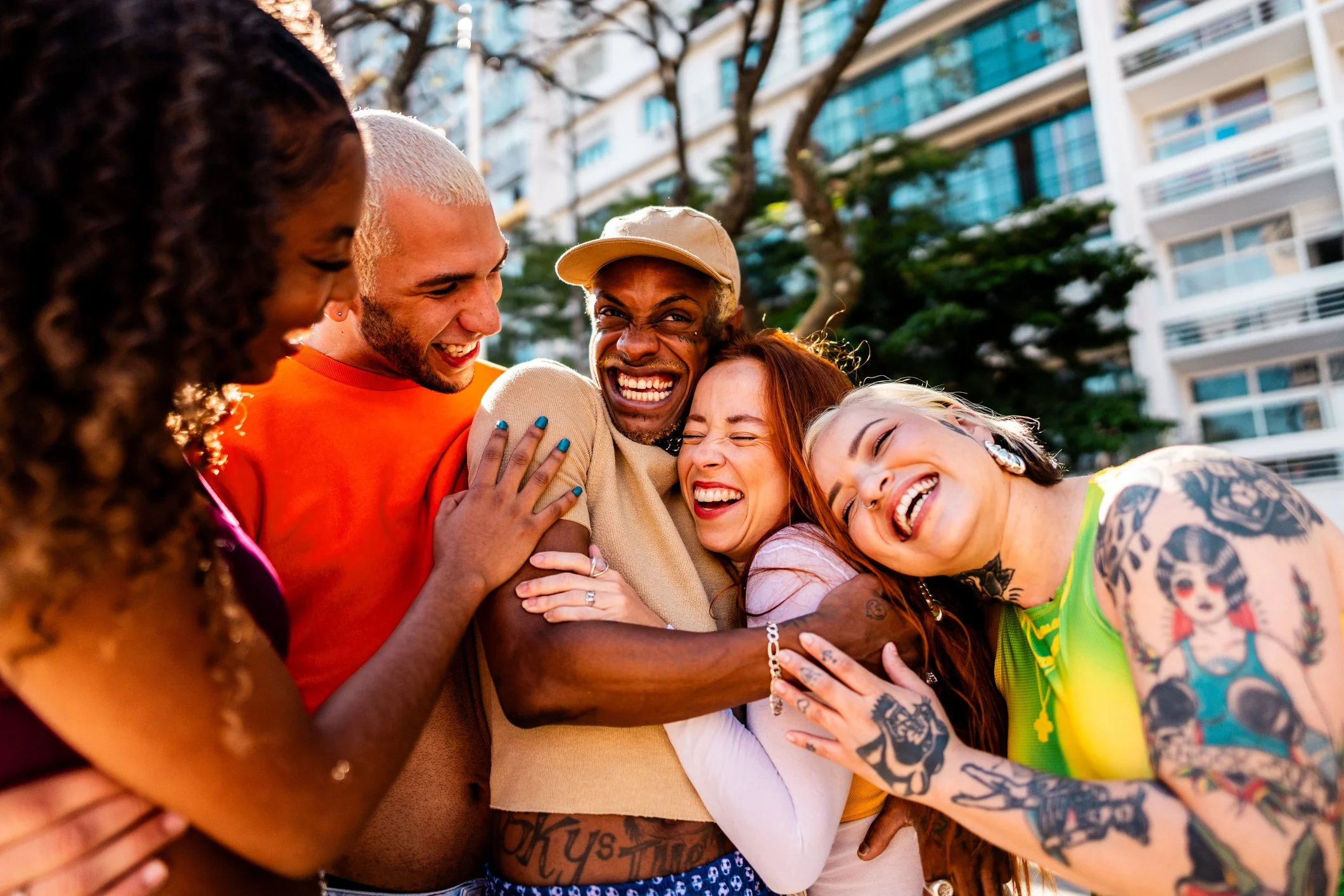 Group of diverse friends smiling and hugging outdoors in an urban setting with trees and apartment buildings in the background.