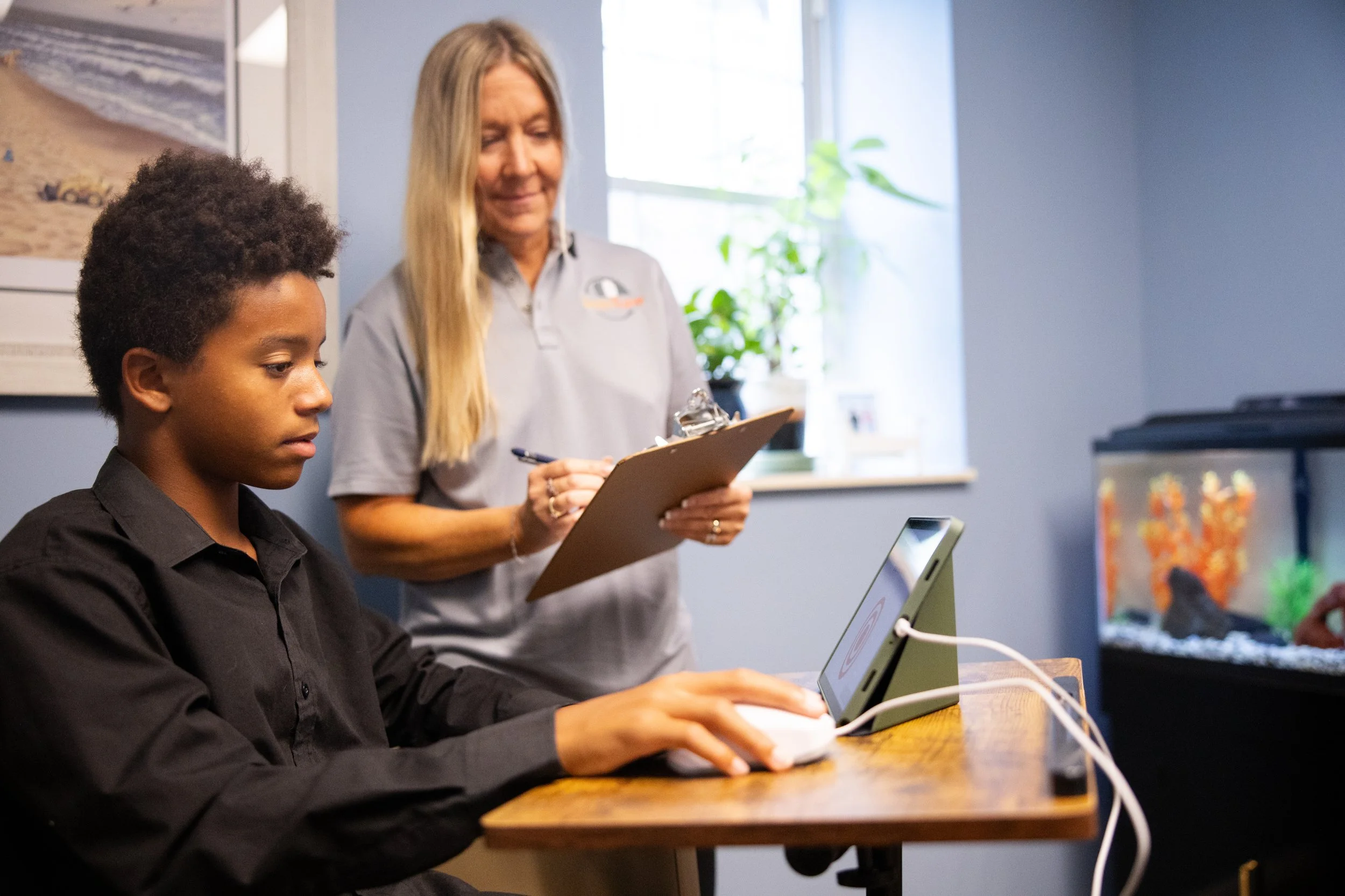 Image of a kid dressed in black doing a Brain Gauge activity on a tablet with a women with a clipboard watching him