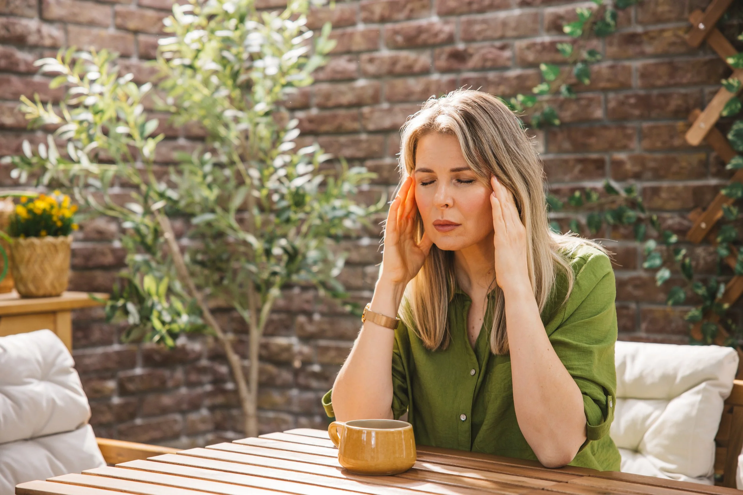 Women in green shirt sitting outside at a table with a coffee mug and her hands on her temples with her eyes closed