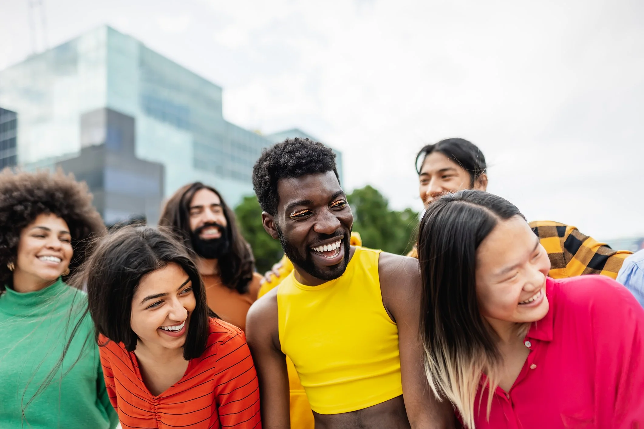 A diverse group of people smiling and hugging outdoors with a modern glass building in the background.