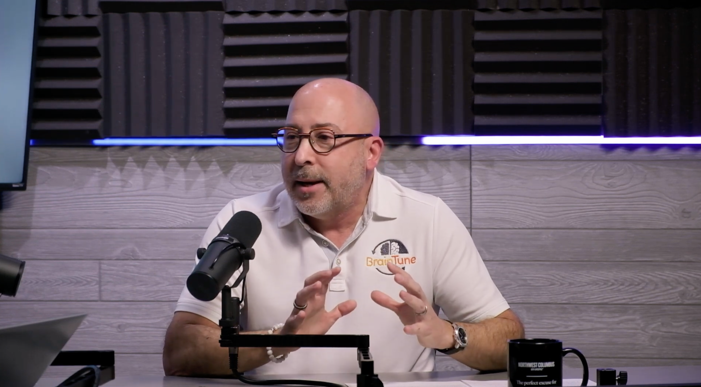 A man wearing glasses and a white polo shirt with a 'BrainTune' logo is speaking into a microphone in a recording studio with foam soundproofing on the walls.