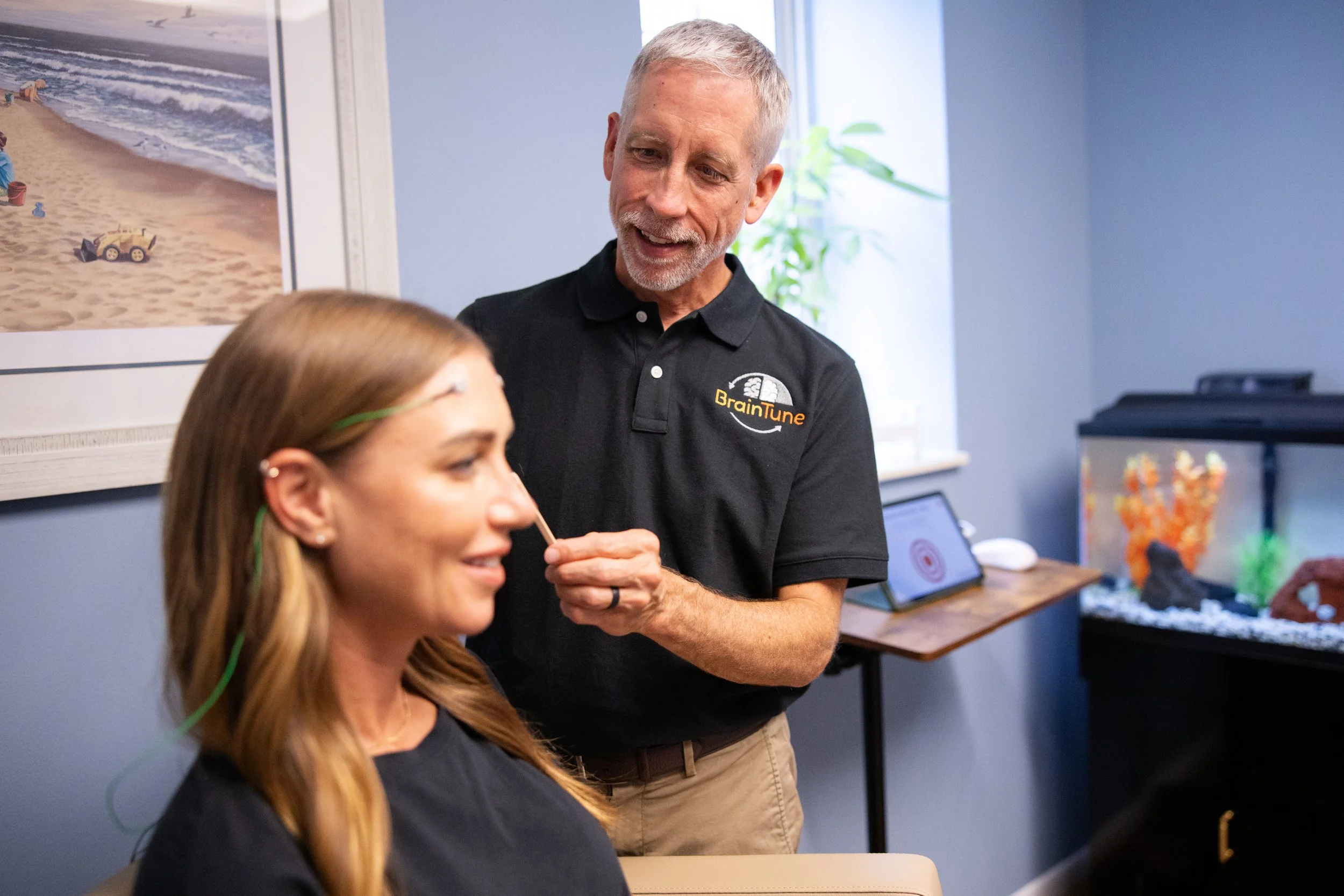 A man in a black shirt with a 'BrainTune' logo is smiling and talking to a seated woman with long blonde hair and glasses while she is receiving a treatment involving small paddles attached to her forehead in a room with light blue walls and a fish tank.