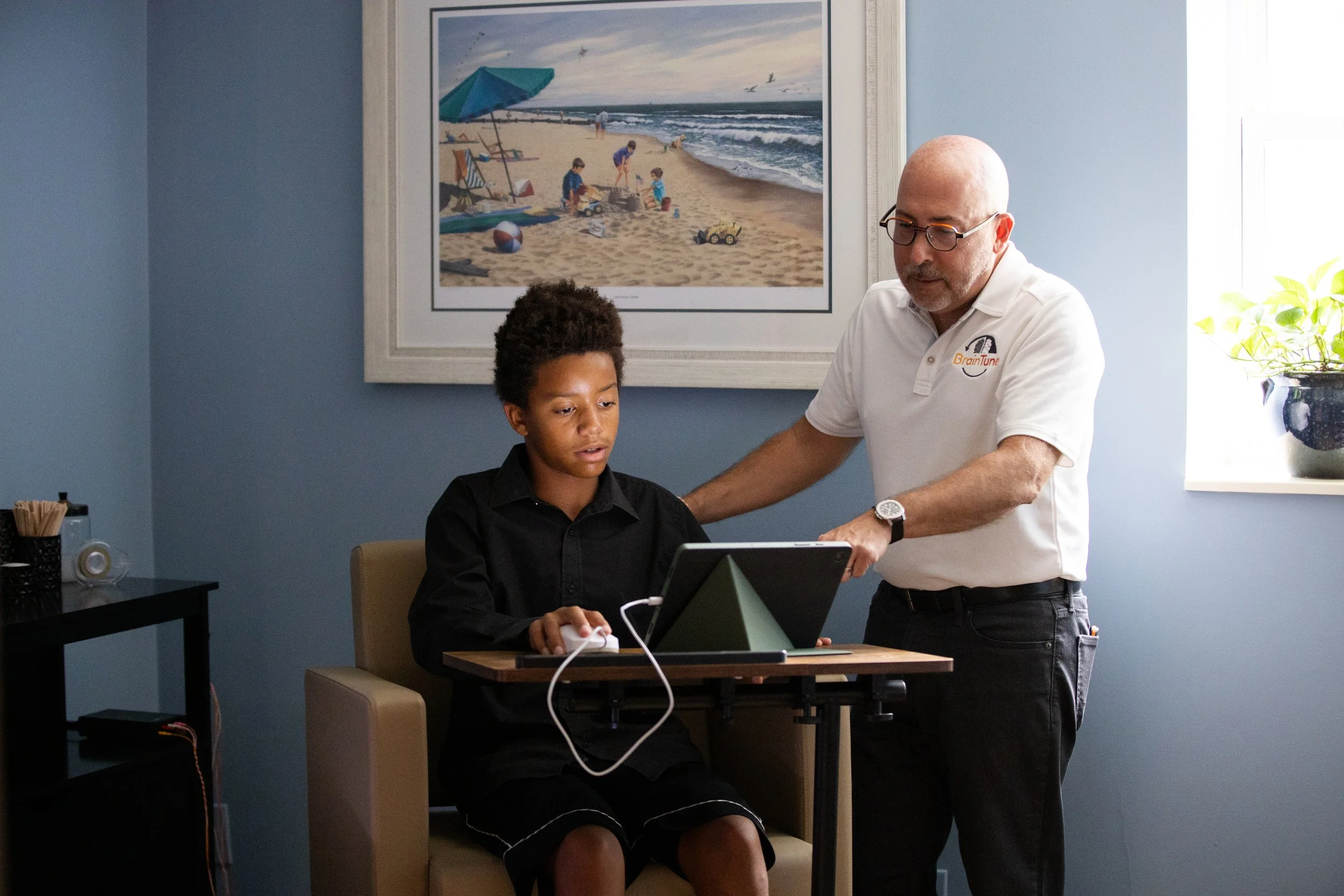 kid in black shirt and pants sitting at a chair with a mini table doing Brain Gauge with an adult in a white shirt standing and pointing to the device.