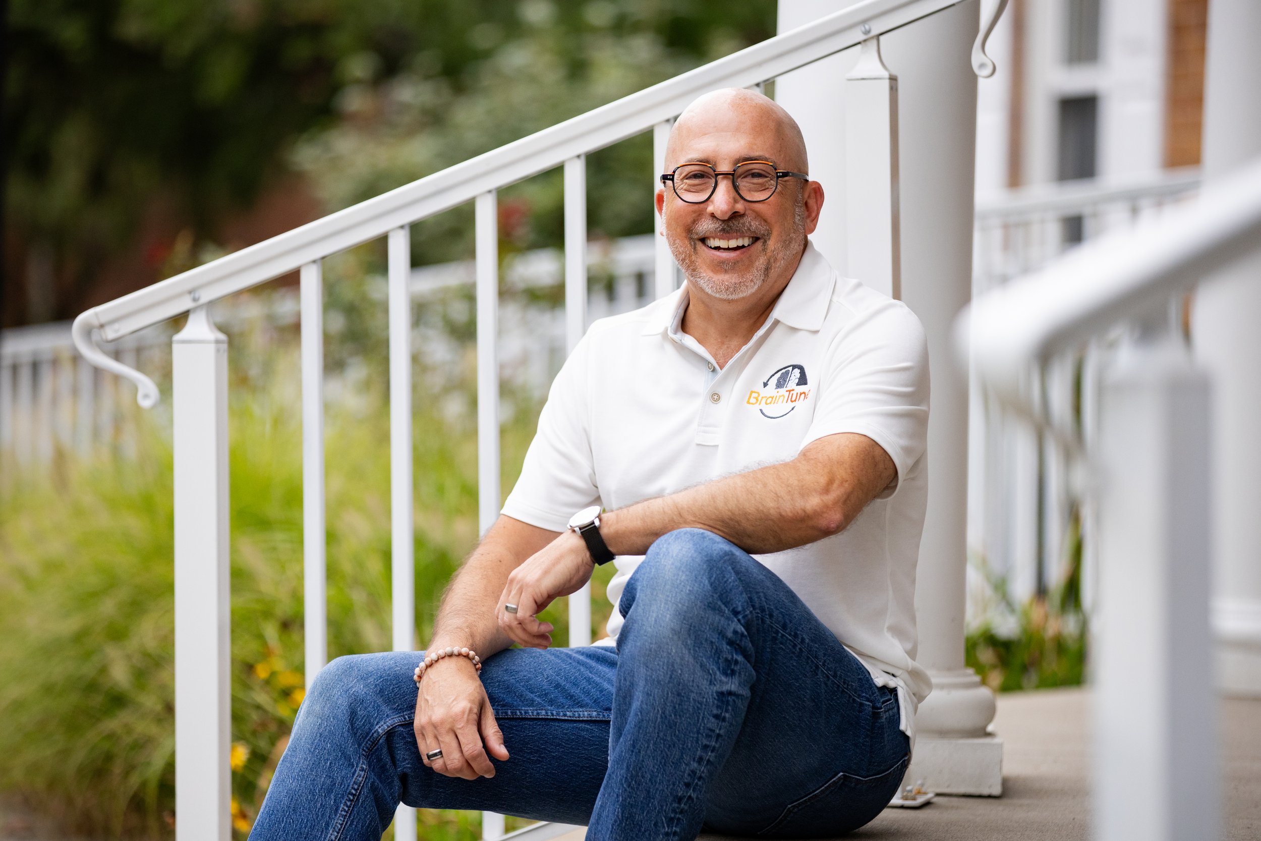 A man in a white shirt and jeans sitting on stairs outside smiling at the camera