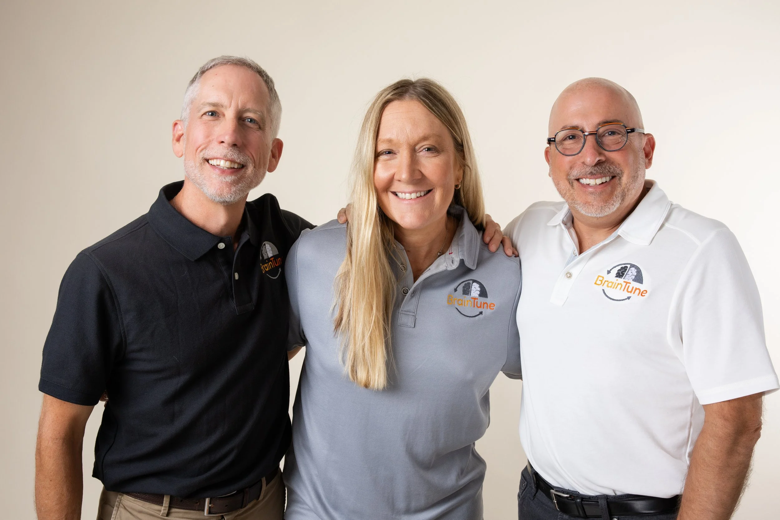 Three people smiling, standing close together, wearing gray and white shirts with the BrainTune logo, against a plain beige background.
