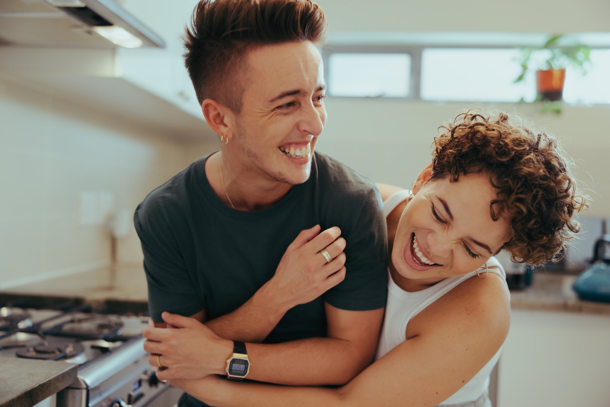 Two women and a man are laughing and enjoying each other's company in a kitchen, with the man and the woman with curly hair leaning close and sharing a joyful moment.