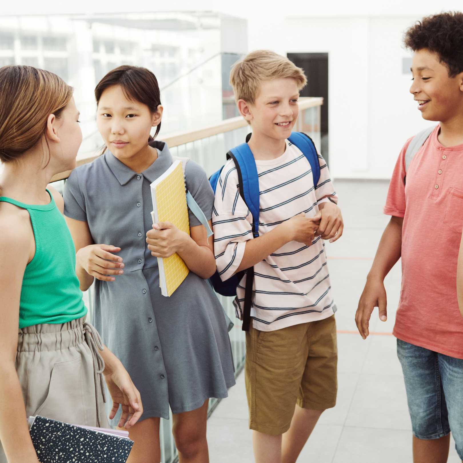 Four children, two boys and two girls, standing and talking in a school hallway, some carrying notebooks and backpacks.
