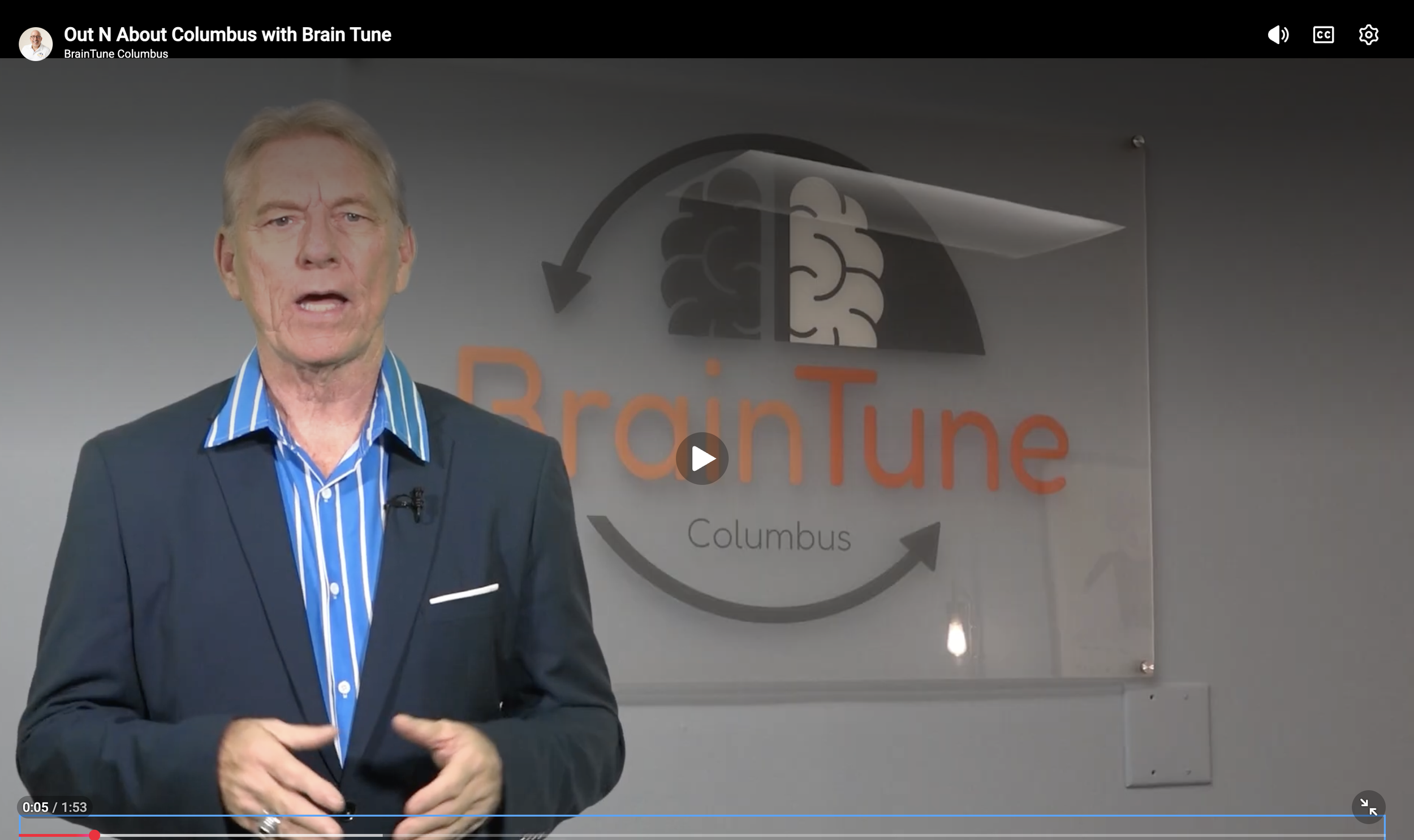 A man in a dark suit and blue striped shirt speaking in front of a wall with a Brain Tune Columbus logo.