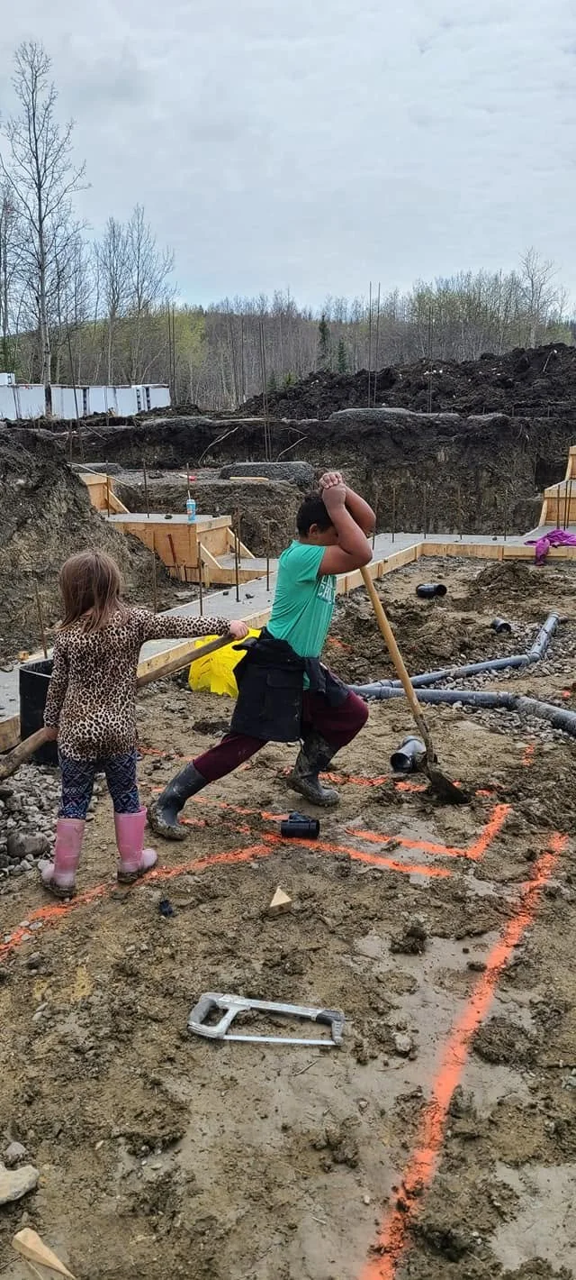 Two children playing with toy construction tools on a muddy construction site, with dirt and pipes, surrounded by excavation areas and trees in the background.