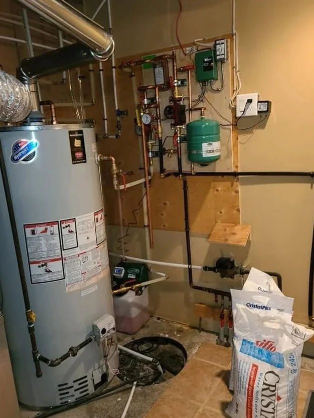 A water heater and plumbing system setup in a basement utility room, with pipes, gauges, and a green expansion tank, along with insulation bags on the floor.
