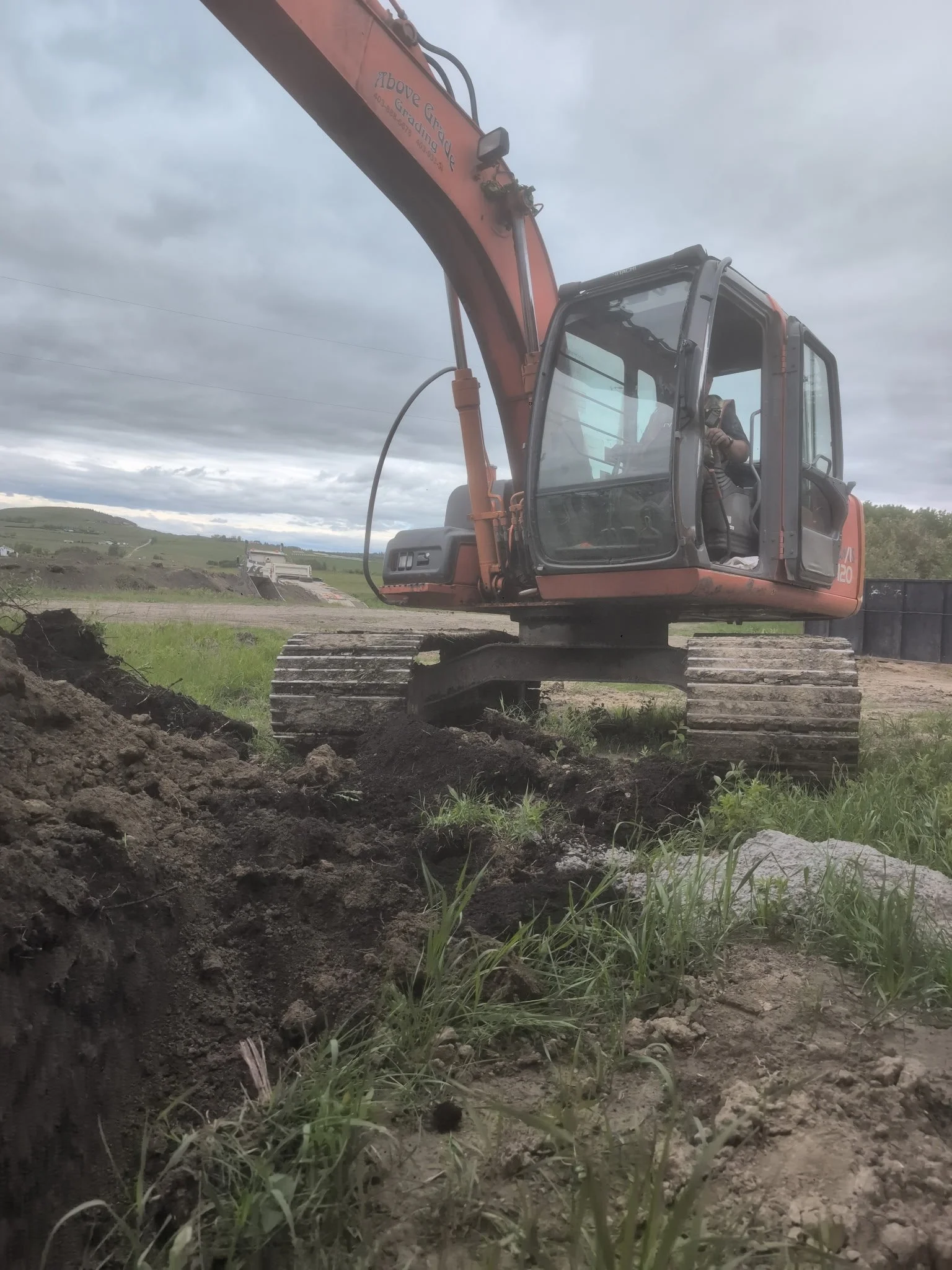 A construction excavator digging into the ground on a rural construction site under a cloudy sky.