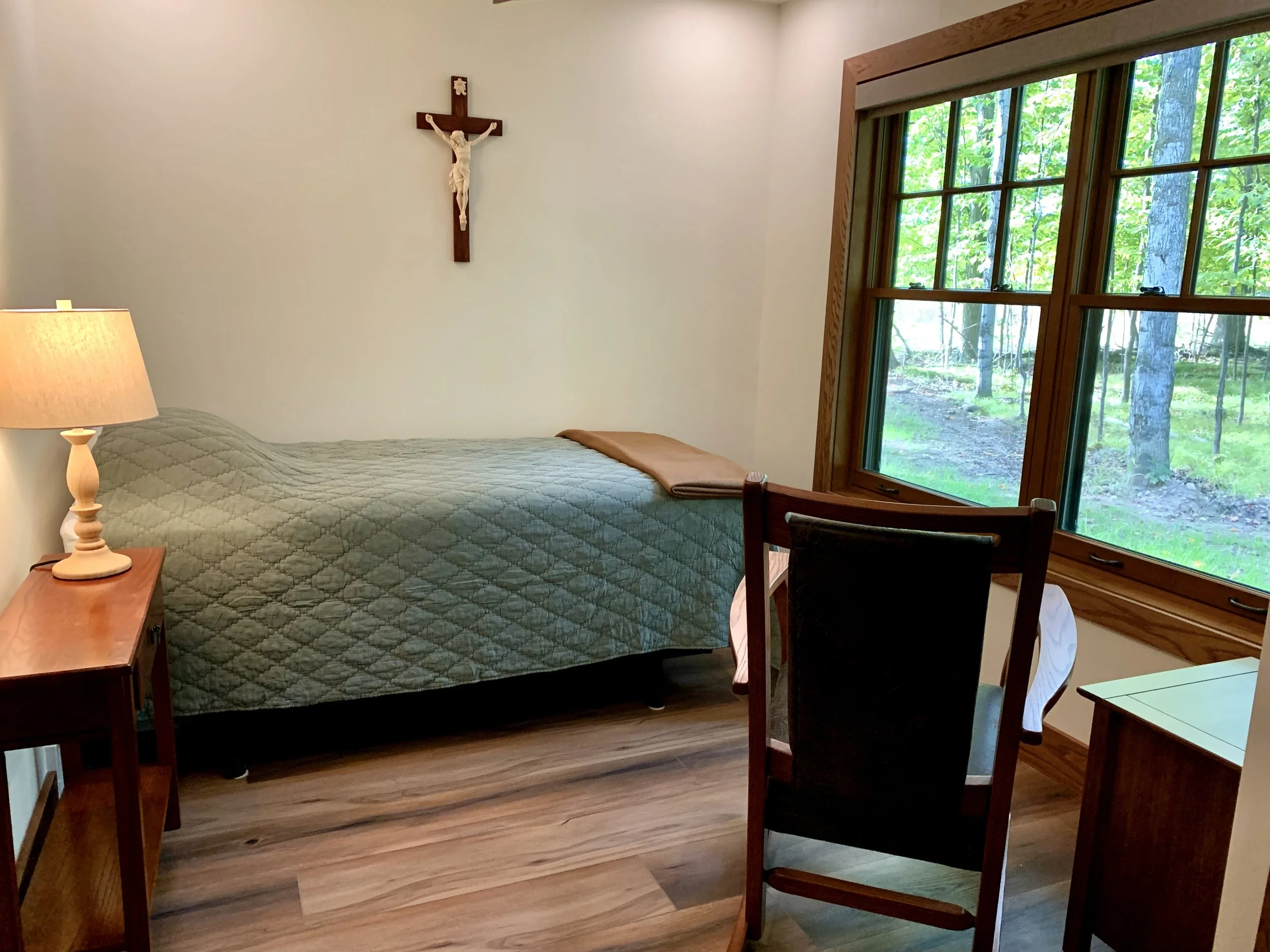 A bedroom with a bed, nightstand, lamp, wooden chair, and window showing trees outside. A crucifix hangs on the wall above the bed.