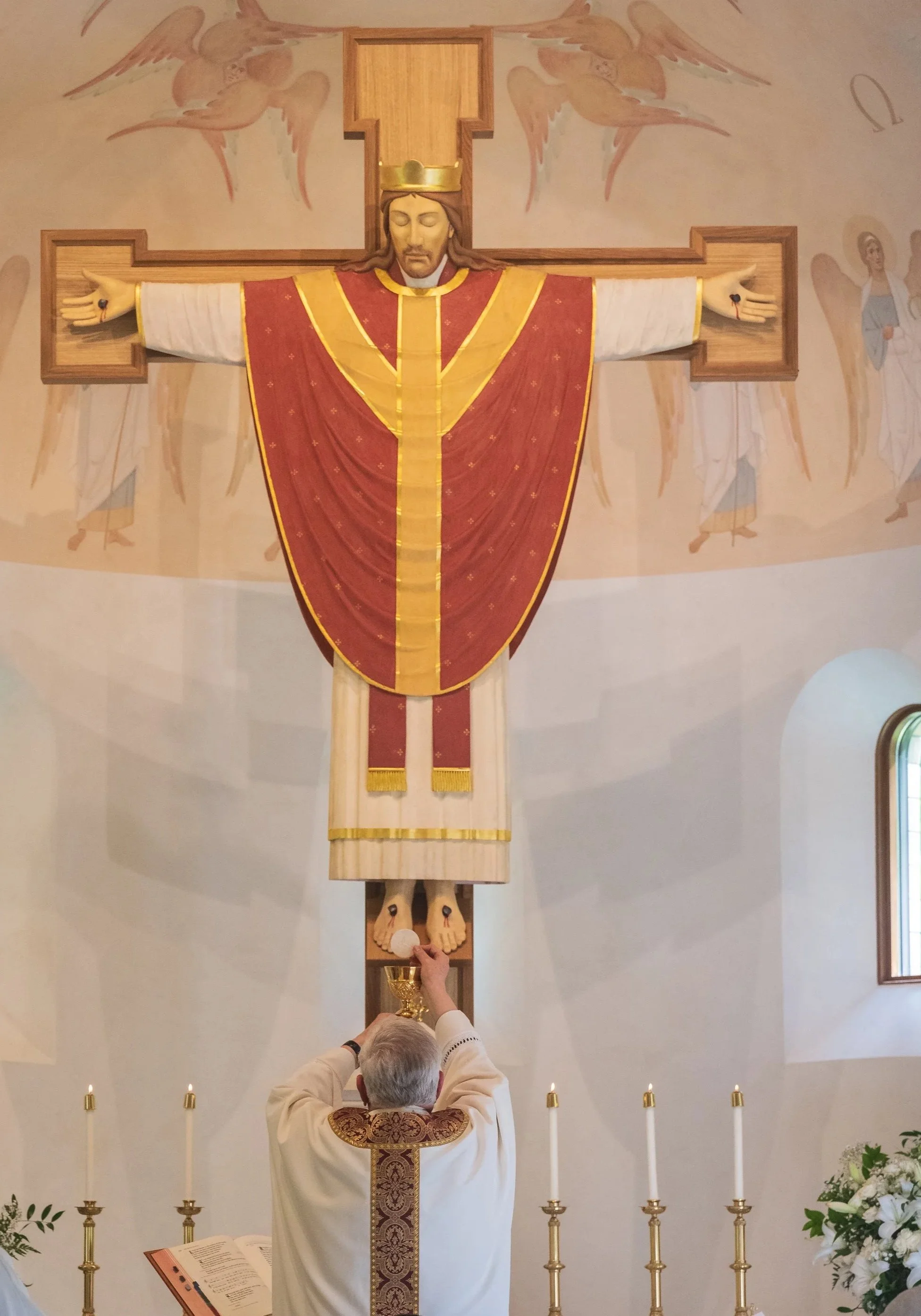 A priest in white robes with gold and red vestments is performing a religious ceremony in front of a large wooden cross with a Jesus figure. The priest is holding a chalice, and the congregation's hand is visible pouring wine into it. Candles and flowers are arranged in front of the altar.