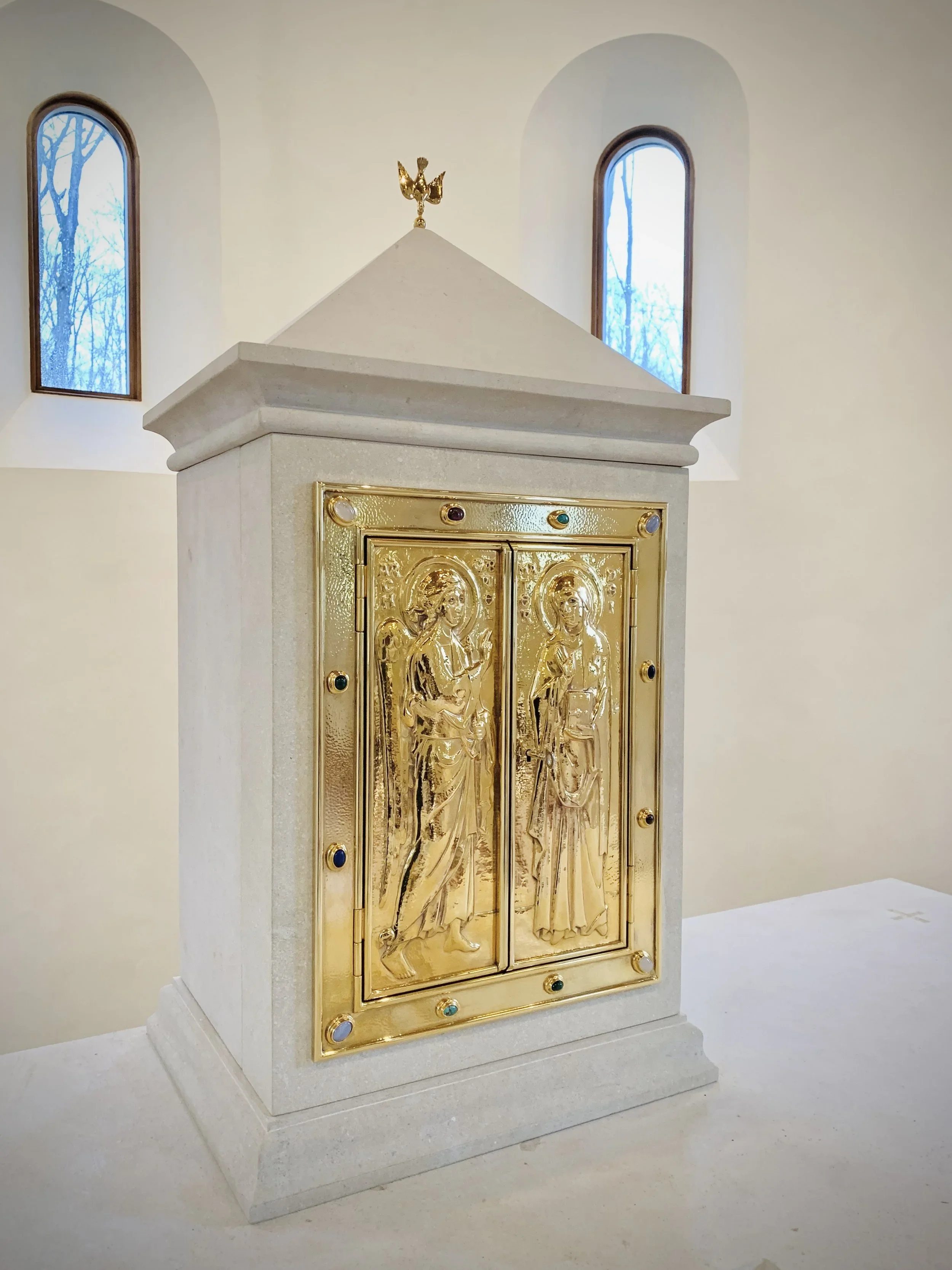 A religious golden altar with engraved images of saints or angels, installed in a room with white walls and two tall arched windows showing a winter scene outside.