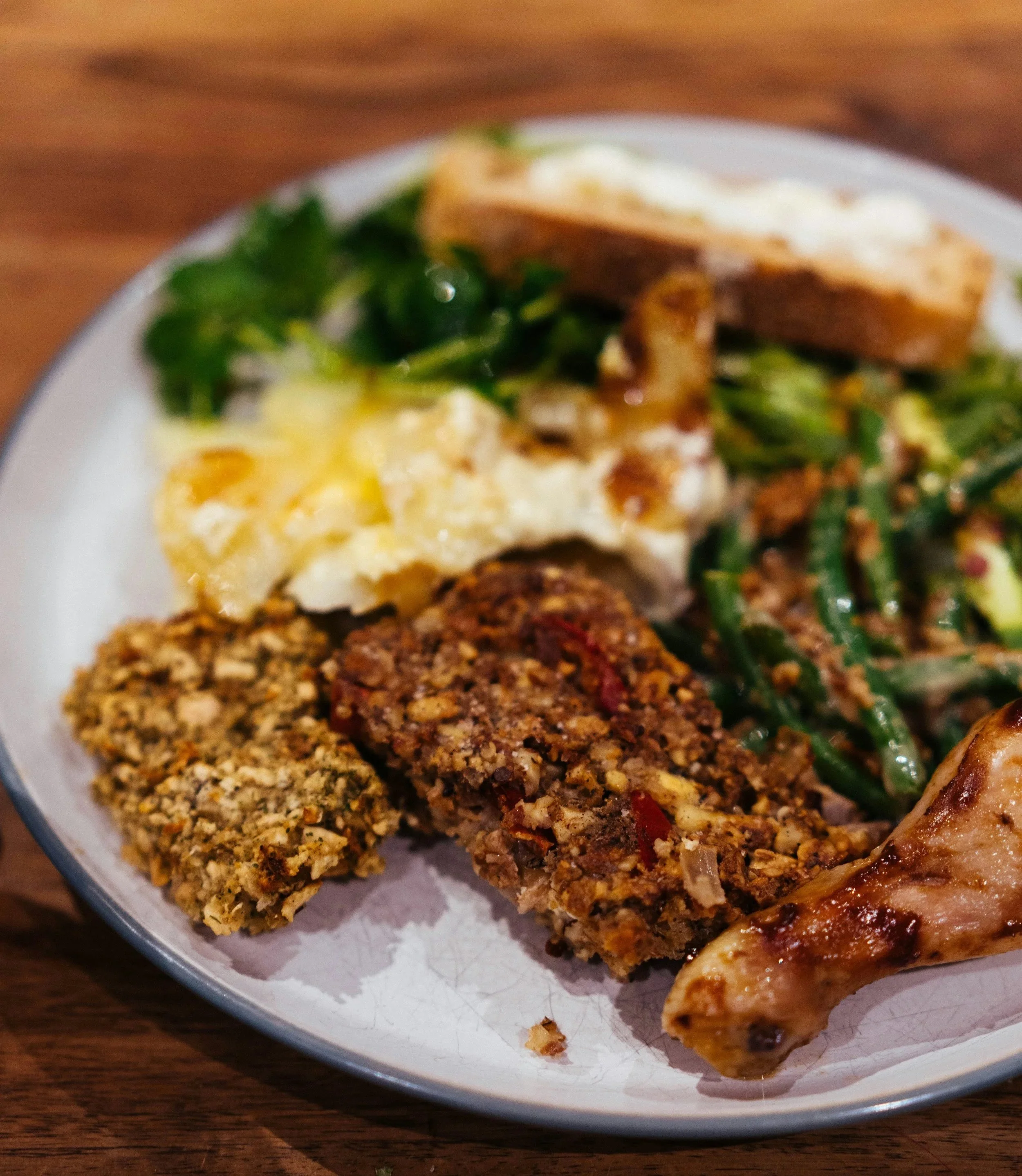 Close-up of a white plate containing seasoned ground meat, cooked chicken drumstick, fried egg, green leafy vegetable, and a side of baked potato with sour cream, on a wooden surface.