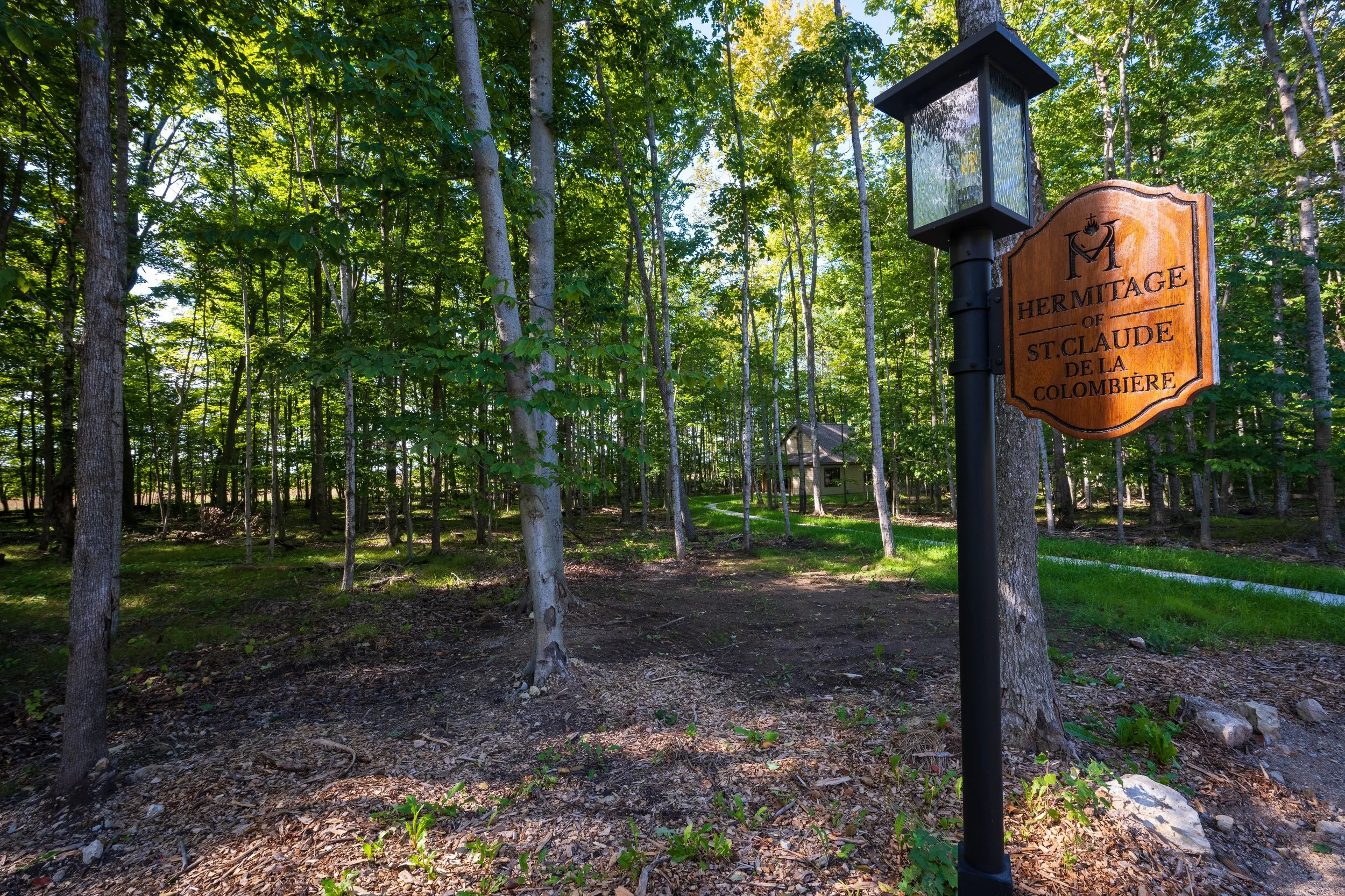 Wooden sign that reads "Hermitage of St. Claude de la Colombière" next to a lamp post, in a wooded area with trees and sunlight filtering through the leaves.