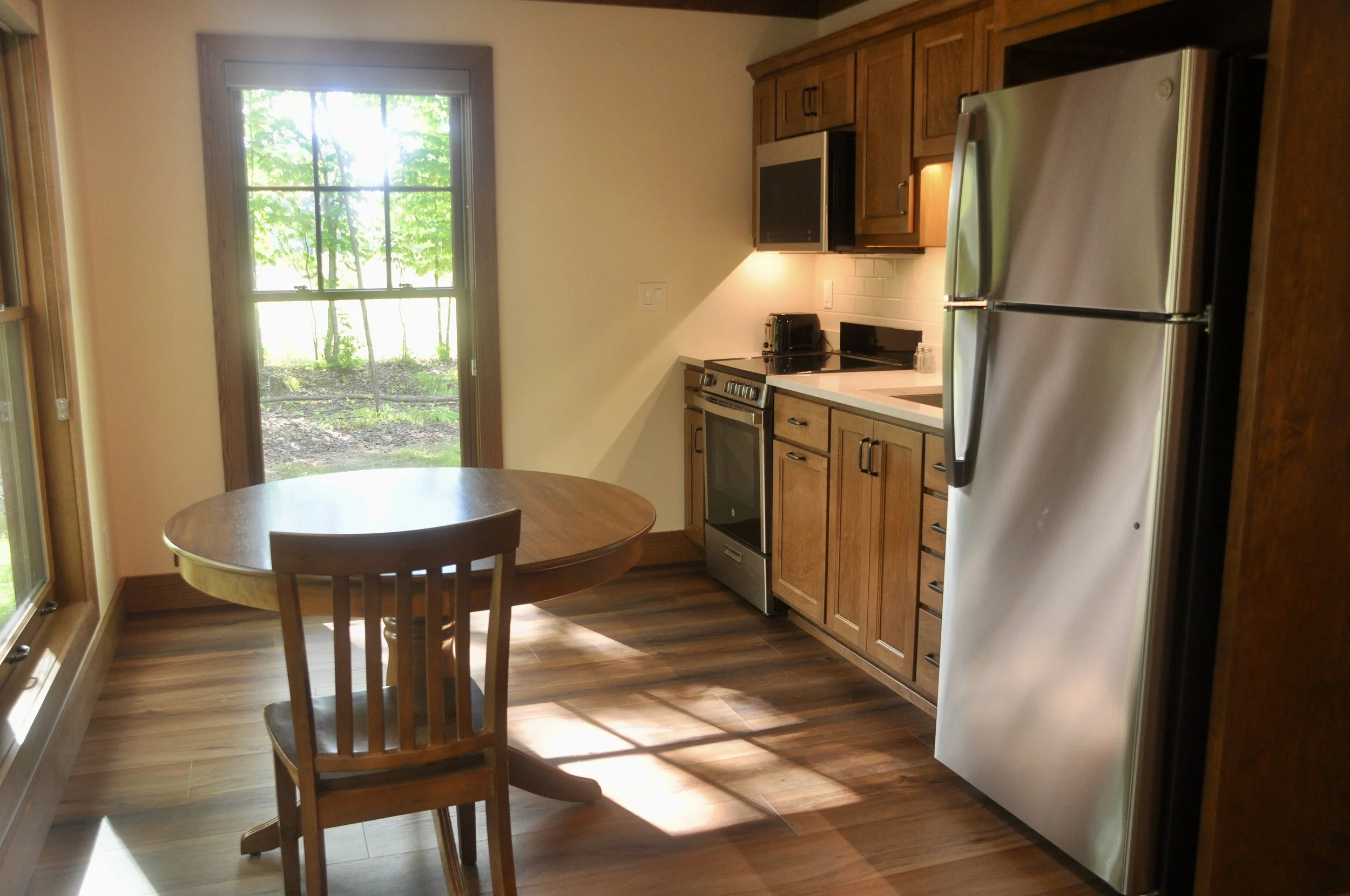 Kitchen with wooden cabinets, stainless steel refrigerator, stove, microwave, and a small round wooden dining table with two chairs. There is a large window showing green trees outside, letting in natural light.