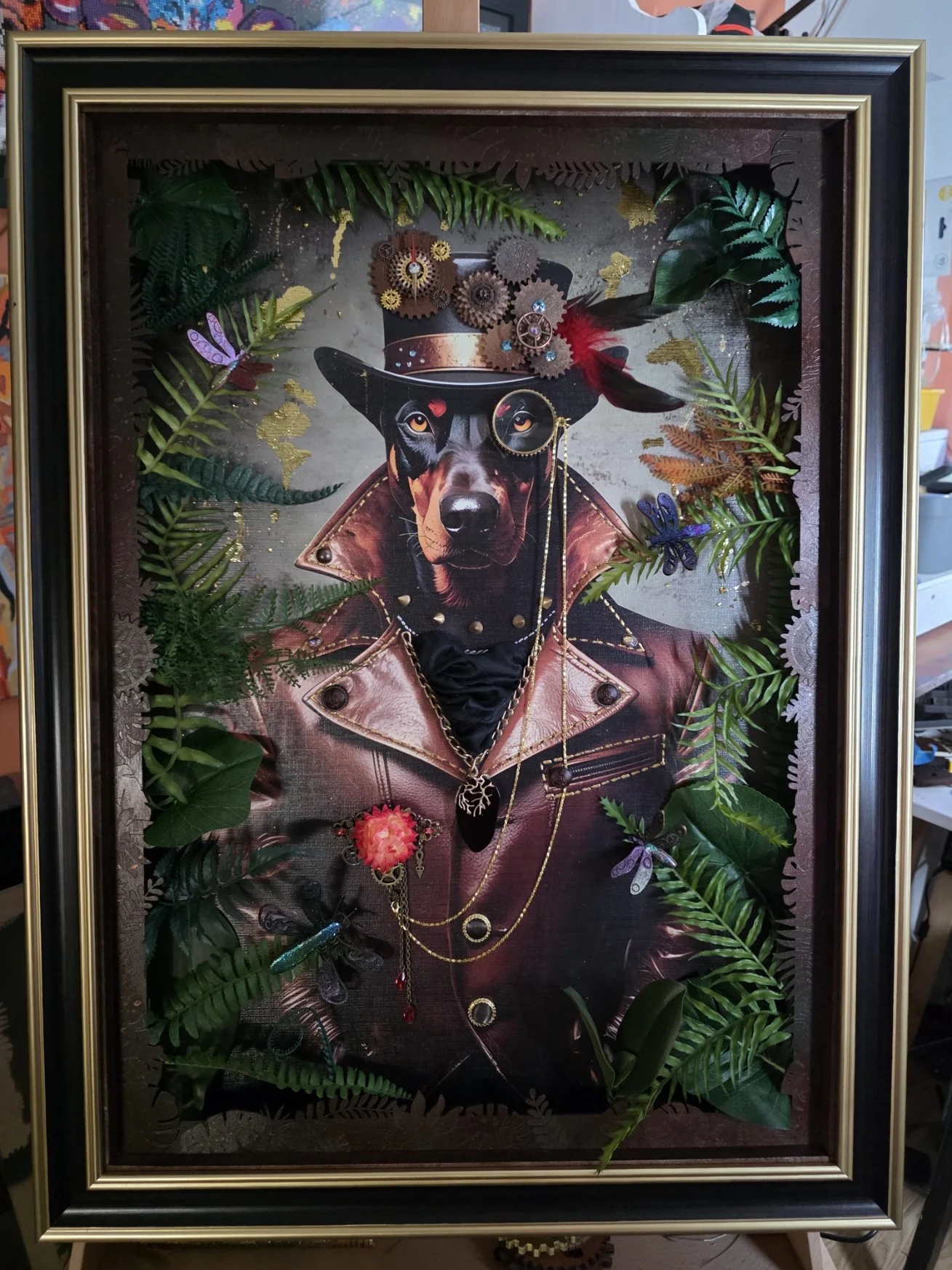 Framed artwork featuring a dog dressed as a cowboy with a black collar, leather jacket, and jewelry surrounded by green leaves and metallic gears and insects.