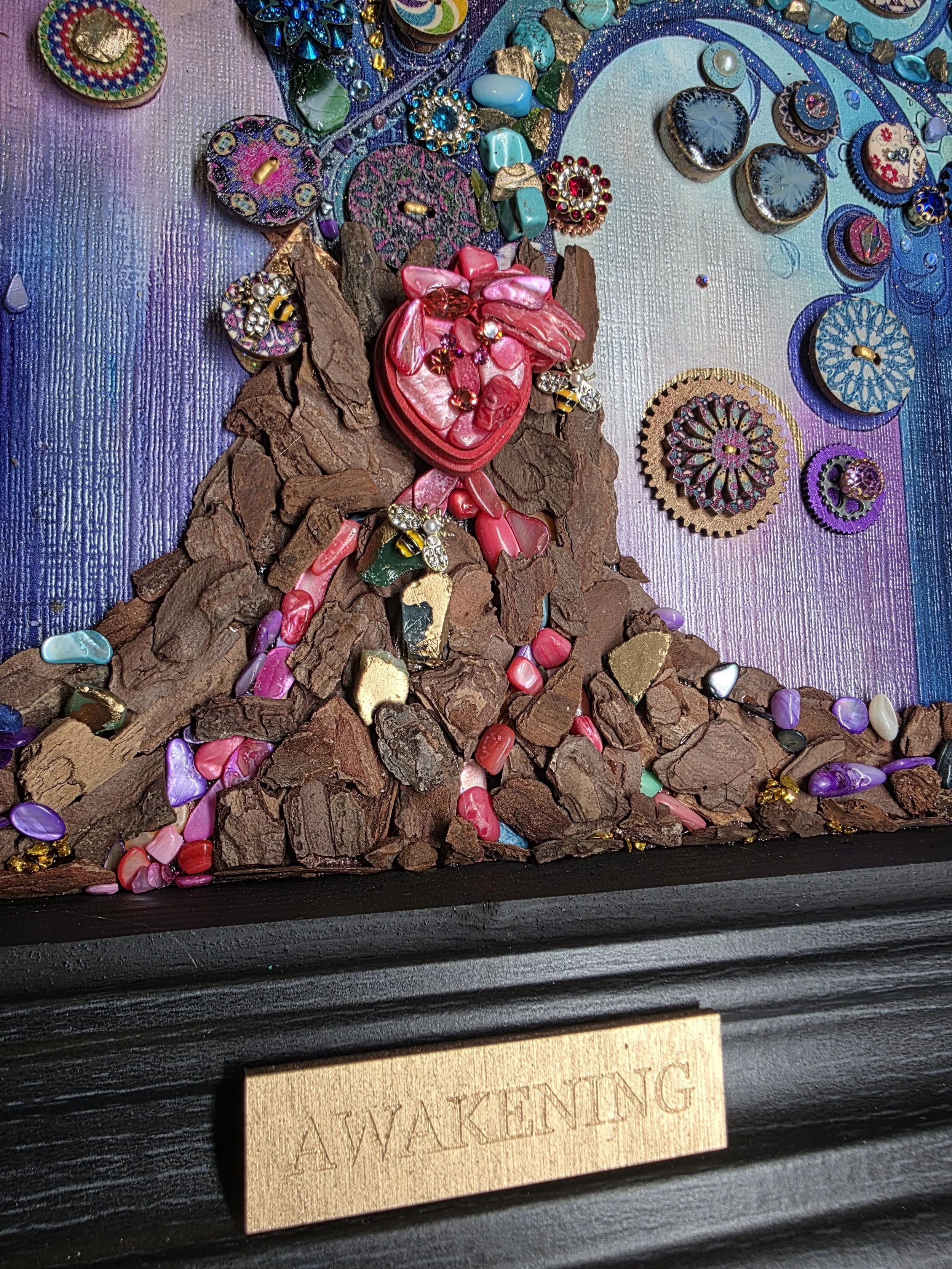A mixed media art piece featuring a representation of a human heart made from pink, red, and green materials, surrounded by various colorful beads, buttons, and decorative objects on a textured background with purple and blue hues. There is a wooden plaque with faint text, partially visible at the bottom.
