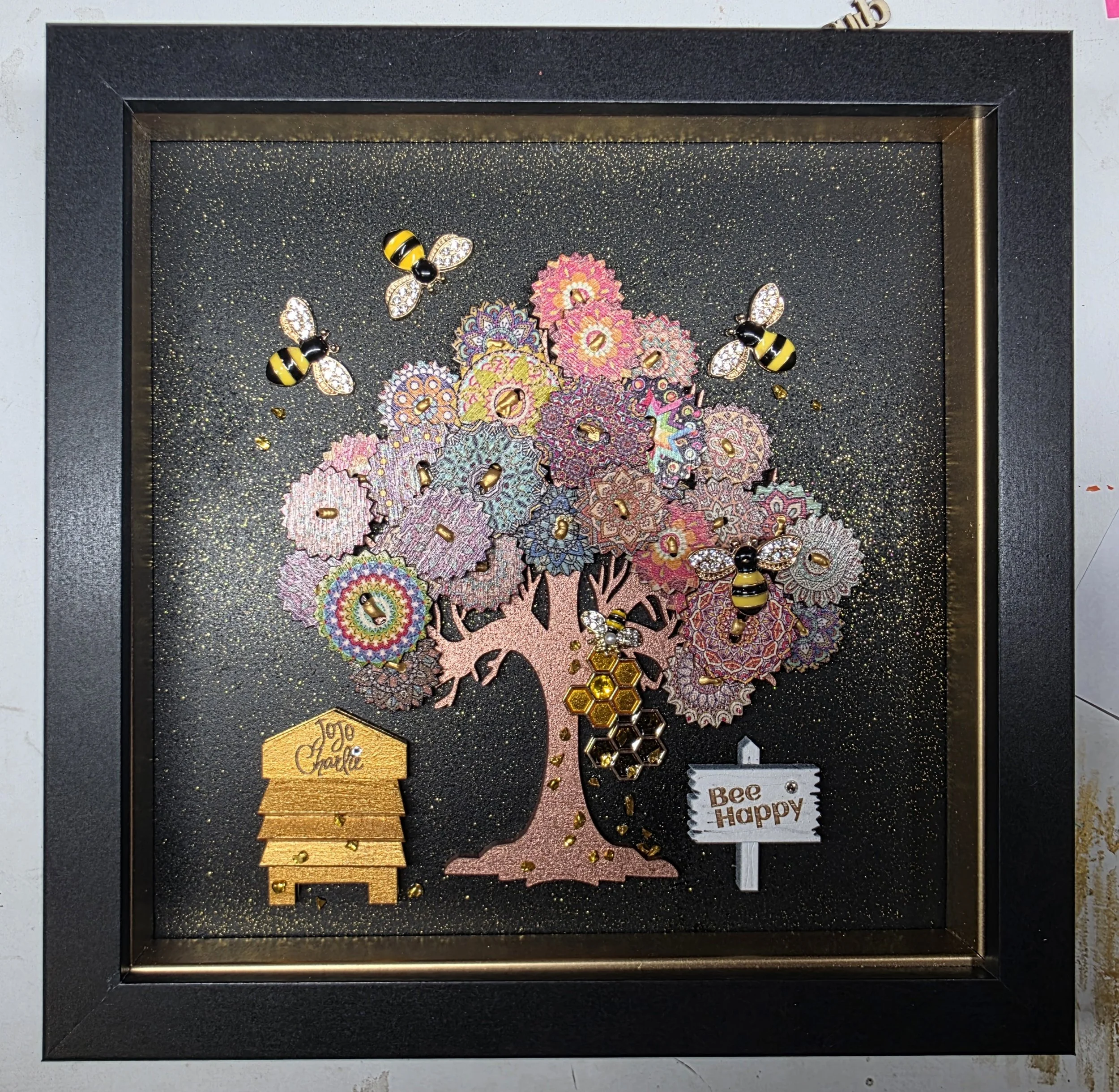 Decorative display case with honeybee-themed jewelry and items, including bee-shaped brooches, necklaces, and stickers, arranged on a glittery black background with a gold-colored honeycomb piece and a small wooden tag that says 'Joy to the World'.